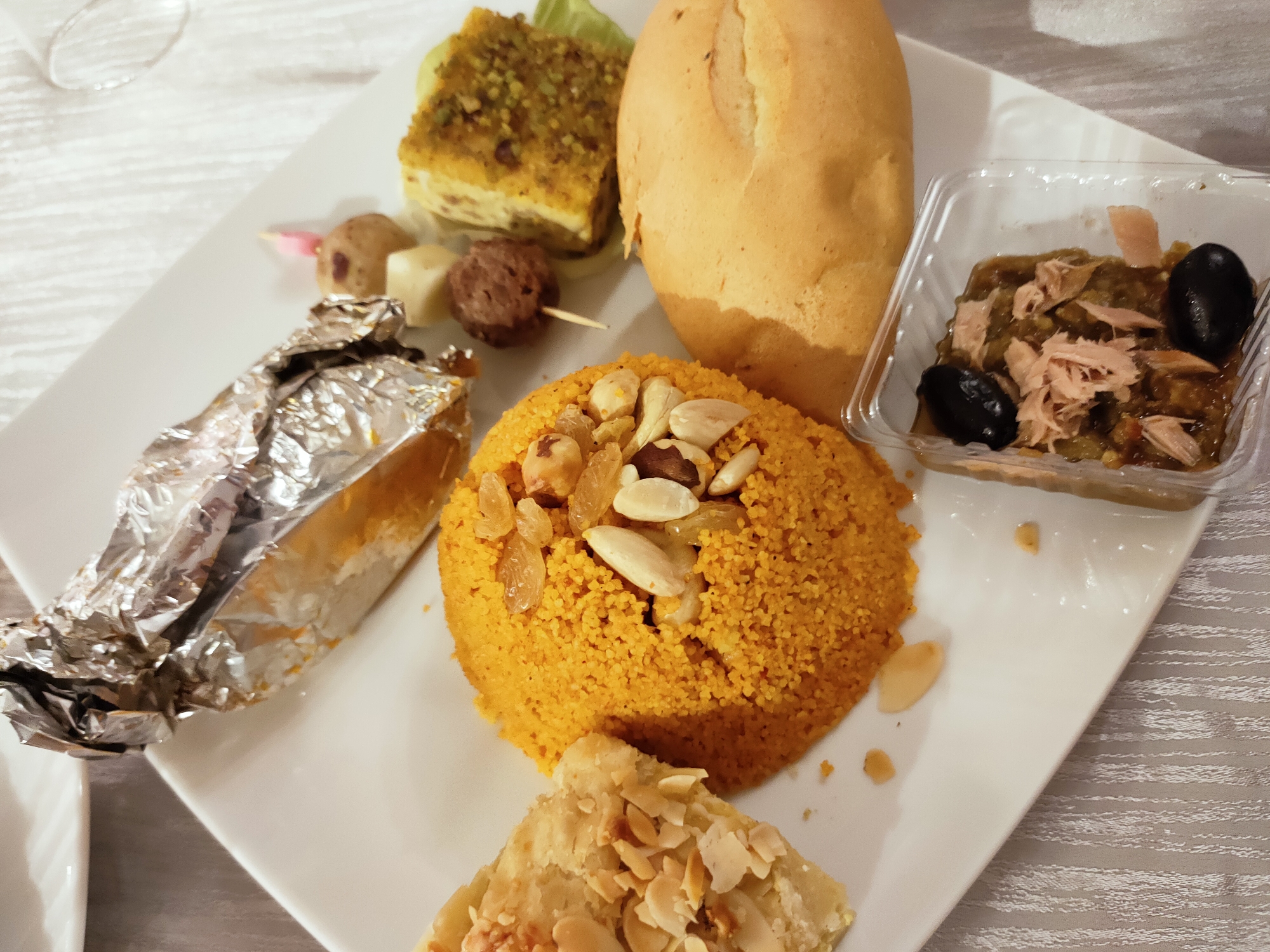 A white square plate holds a variety of food items on a table with a patterned tablecloth. In the upper left quadrant, a glass with a rim is visible, partially out of focus. Directly to its right, a rectangular piece of food topped with green crumbs sits on a lettuce leaf. To the right of this is a light brown, oval-shaped bread roll. Below the bread roll and to the right is a small, clear plastic container with a green, chunky mixture inside, topped with shredded pinkish meat and two black olives.
Moving towards the center of the plate, a skewer with small, rounded food items is visible. These include a dark brown meatball, a white cube, and a pinkish, cone-shaped item. To the left of the skewer is a portion of food wrapped in crinkled aluminum foil.
Dominating the center of the plate is a mound of bright orange, granular food, likely couscous, adorned with almonds and raisins. Below this orange mound, a triangular piece of pastry topped with sliced almonds is situated. The overall arrangement suggests a sampling of different dishes, possibly a tasting menu or a variety platter. The lighting is dim, creating some shadows and highlighting the textures of the food. There are no people visible in the image. The scene appears to be an indoor setting, likely a restaurant or dining table.