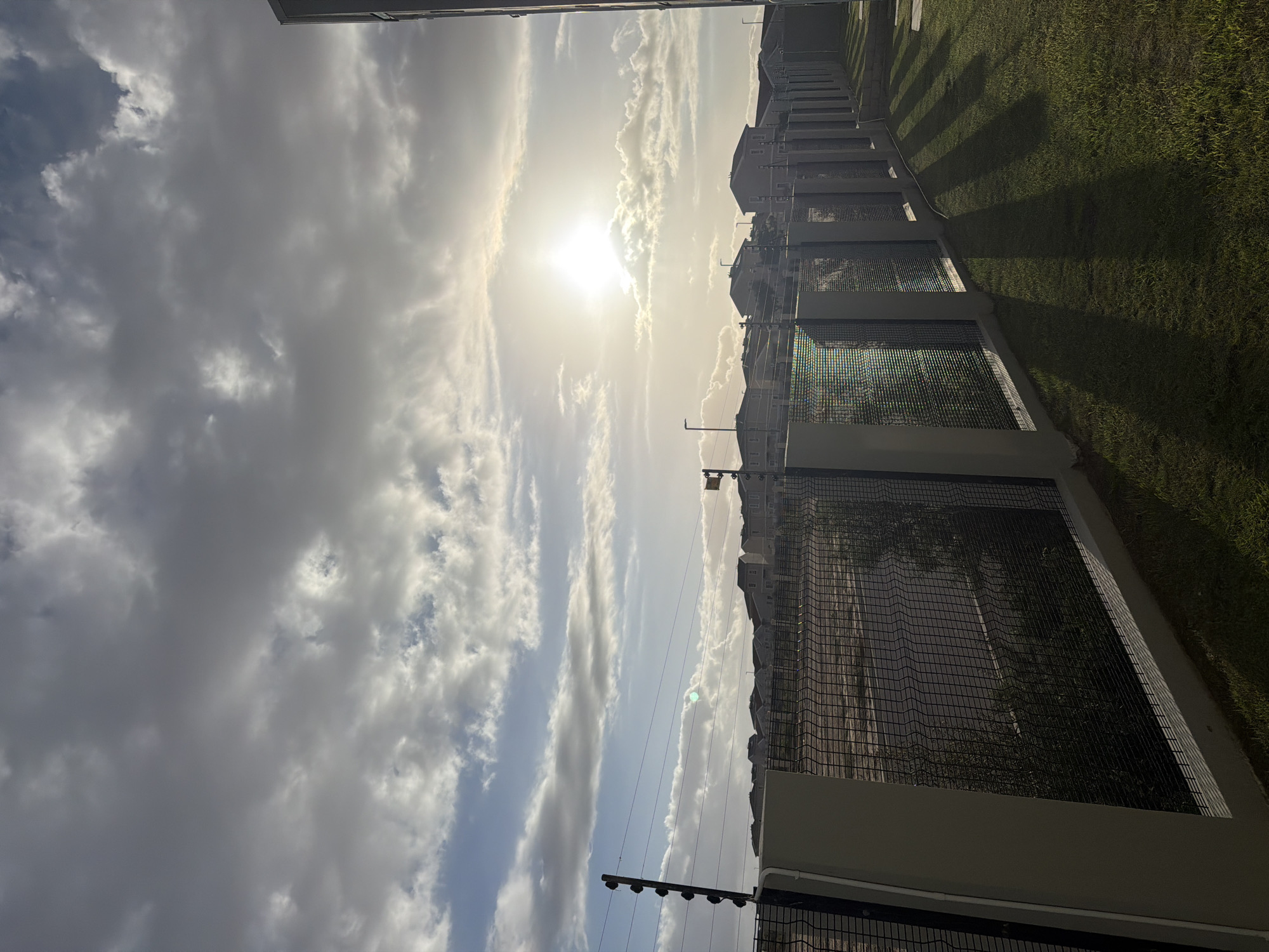 The image, when viewed in its correct orientation, captures an outdoor scene within what appears to be a residential complex, likely in Cape Town, South Africa. The time of day is characterized by a low, bright sun, indicating either early morning or late afternoon, as evidenced by the long, distinct shadows cast across the foreground.
The sky occupies a significant portion of the upper frame, featuring a mix of white and grey clouds against a light blue backdrop. The sun is a brilliant, intense light source, appearing as a bright disc partially veiled by clouds, creating a strong lens flare effect and illuminating the scene with a golden hue.
On the right side of the image, a series of multi-story residential buildings or houses are visible. These structures exhibit a uniform architectural style with light-colored (grey or off-white) facades and darker upper sections or roofs. They are set behind a substantial security fence line.
The foreground is dominated by a neatly maintained expanse of green grass. Running alongside this grassy area and separating it from the buildings is a tall, dark, mesh-like security fence with concrete bases. Evidence of an electric fence system, including wires and insulators, is visible along the top of the fence. The strong sunlight casts prominent, parallel shadows from these fences and possibly unseen structures onto the grassy ground.
No people or specific activities are depicted in the scene. There is no visible text in the image. The overall impression is one of a well-secured and well-maintained residential area under clear, sunny, yet partly cloudy skies.