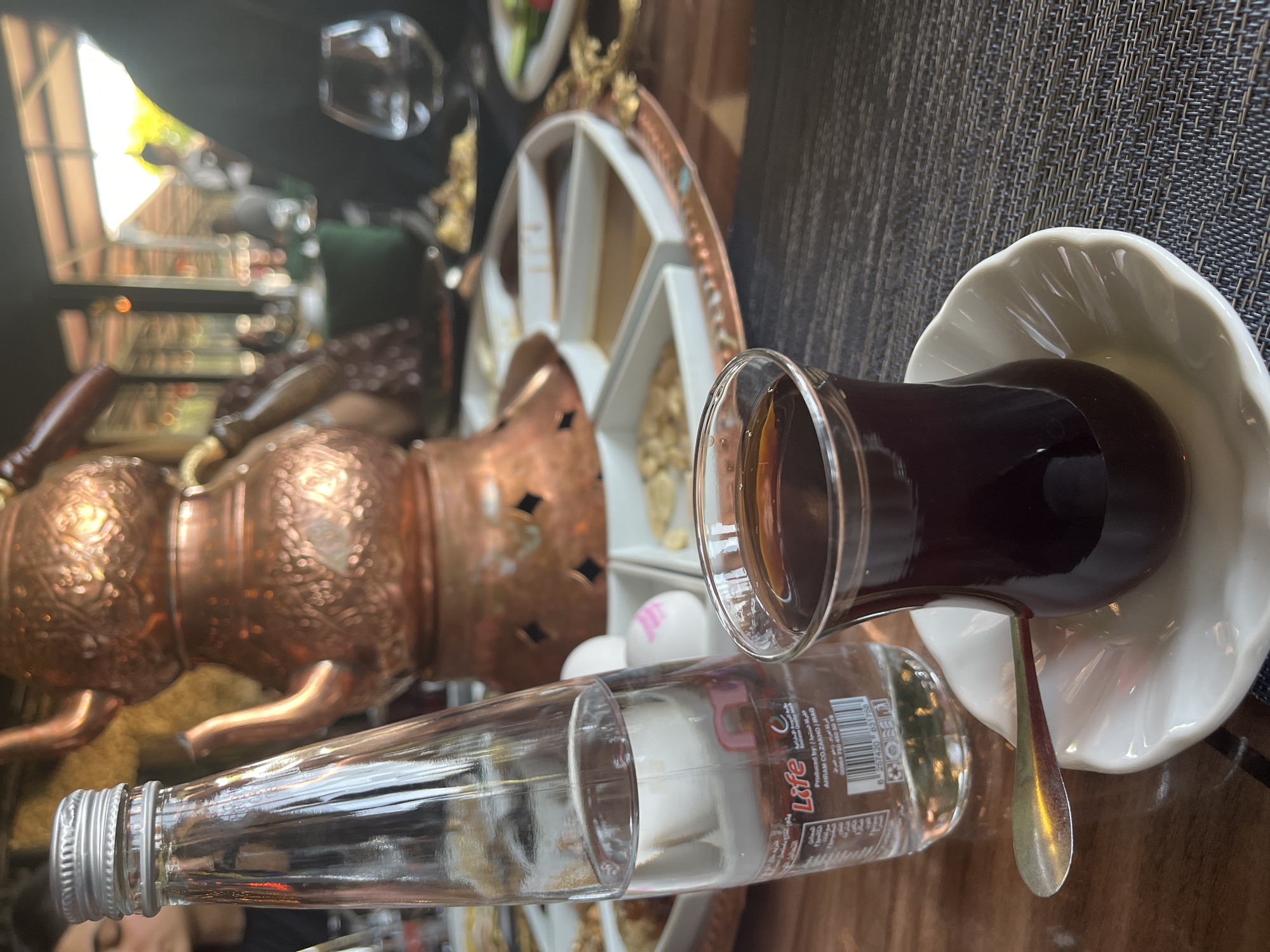 A close-up, eye-level, medium shot captures a table setting at an outdoor restaurant with a blurred background. In the foreground, a clear glass of dark liquid, likely tea, sits on a white saucer with a scalloped edge. A small silver spoon rests on the saucer, angled towards the glass. To the left, a partially visible glass bottle of water, with a silver cap, stands upright. The bottle has a label with text and barcodes. Behind the bottle and glass, a traditional copper tea kettle, intricately engraved, is prominently featured. A round copper tray holds multiple white ceramic dishes filled with various food items, possibly nuts and dips, arranged around the kettle. In the blurred background, there are glimpses of other diners, outdoor seating, and greenery, suggesting a sunny day. A person's head and shoulders are visible on the far left, and another person's dark clothing is visible on the right. The overall impression is a relaxed dining experience in an open-air environment.