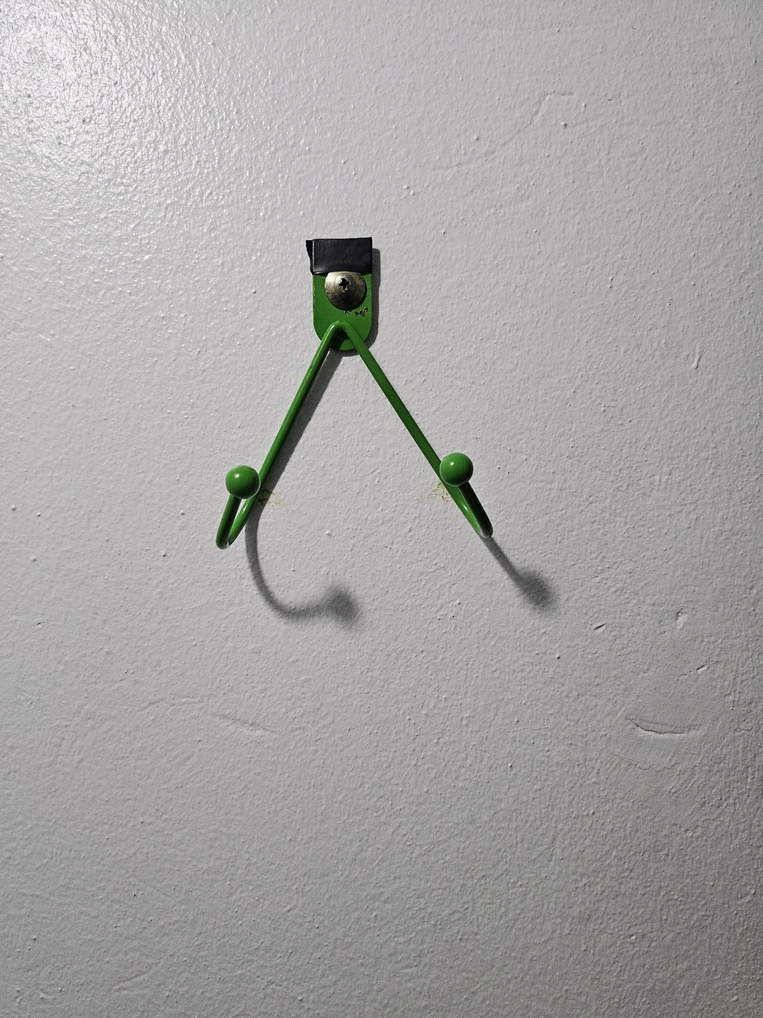 A green metal hook is mounted on a white textured wall. The hook has two prongs, each ending in a small green ball. A black piece of tape is visible at the top of the hook, covering part of the screw that attaches it to the wall. The wall is illuminated from the left, casting shadows from the hook onto the wall. The scene is indoors and appears to be in Batna, Algeria.
