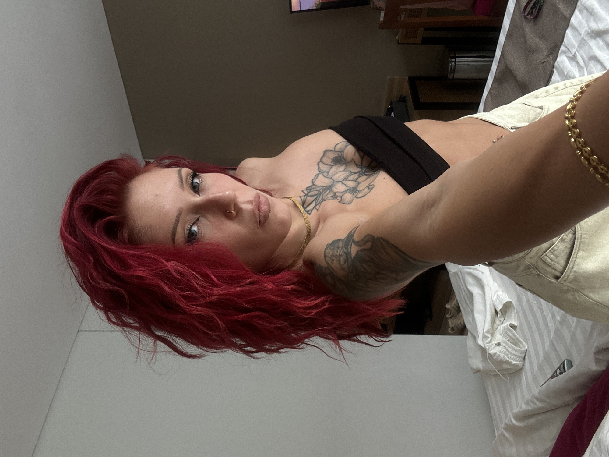 A fair-skinned woman with bright red curly hair lies on a white bed. She wears a black top and light-colored shorts, and has a gold chain necklace and bracelet. Tattoos are visible on her chest and arm. The background includes a wall and a blurred interior with furniture. The location and specific city in Indonesia are not discernible from the image.