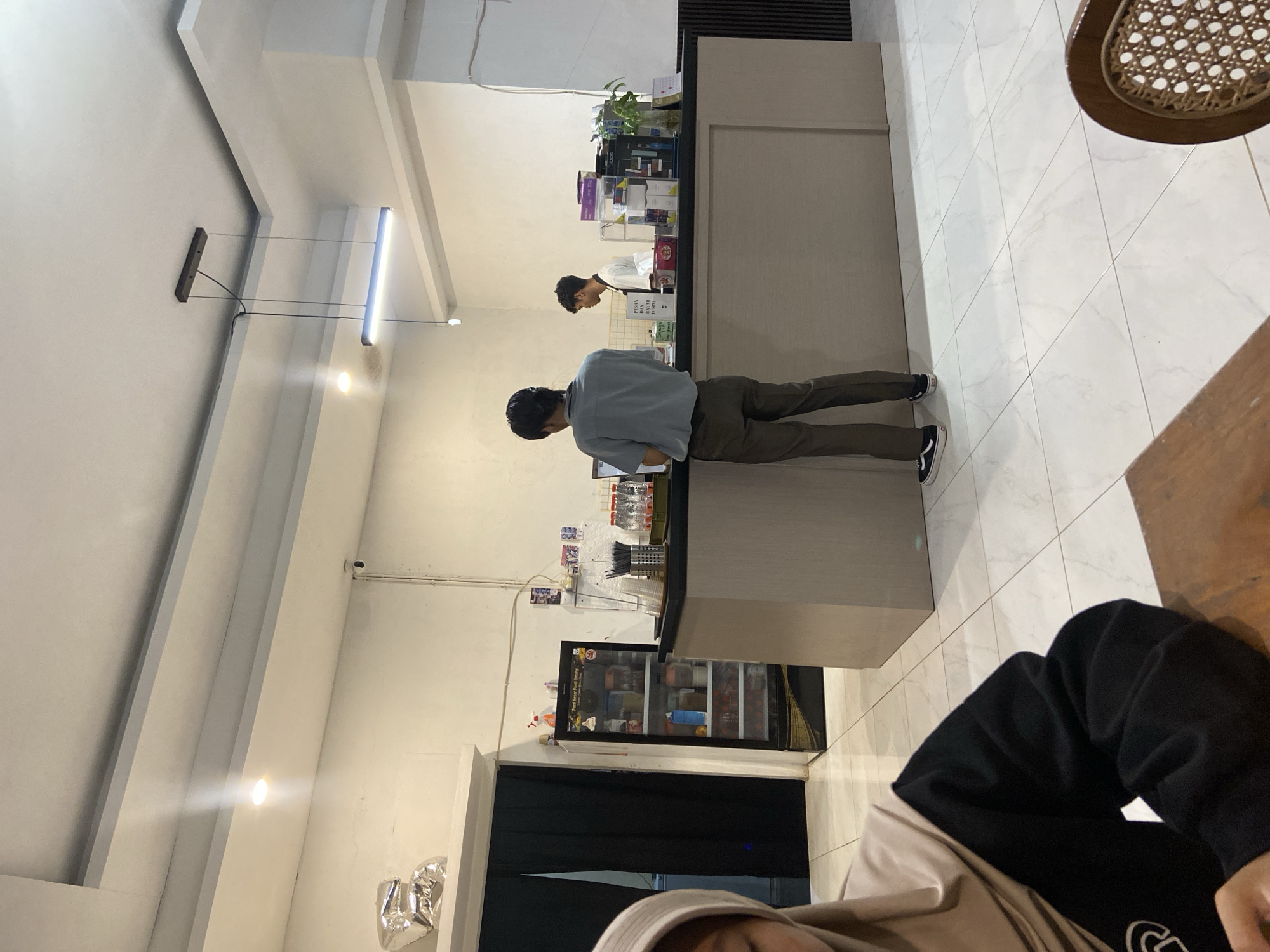 In Sekar Mawar, Indonesia, a commercial establishment features a counter with two employees and a customer. The counter area displays various items, including beverages, snacks, and electronic smoking devices. A refrigerator unit is positioned to the left of the counter, stocked with bottled drinks. The floor is tiled with a marble pattern, and the ceiling is illuminated by recessed lighting and a linear pendant fixture. A person in the foreground, partially visible, is seated at a table, suggesting a dining or waiting area.