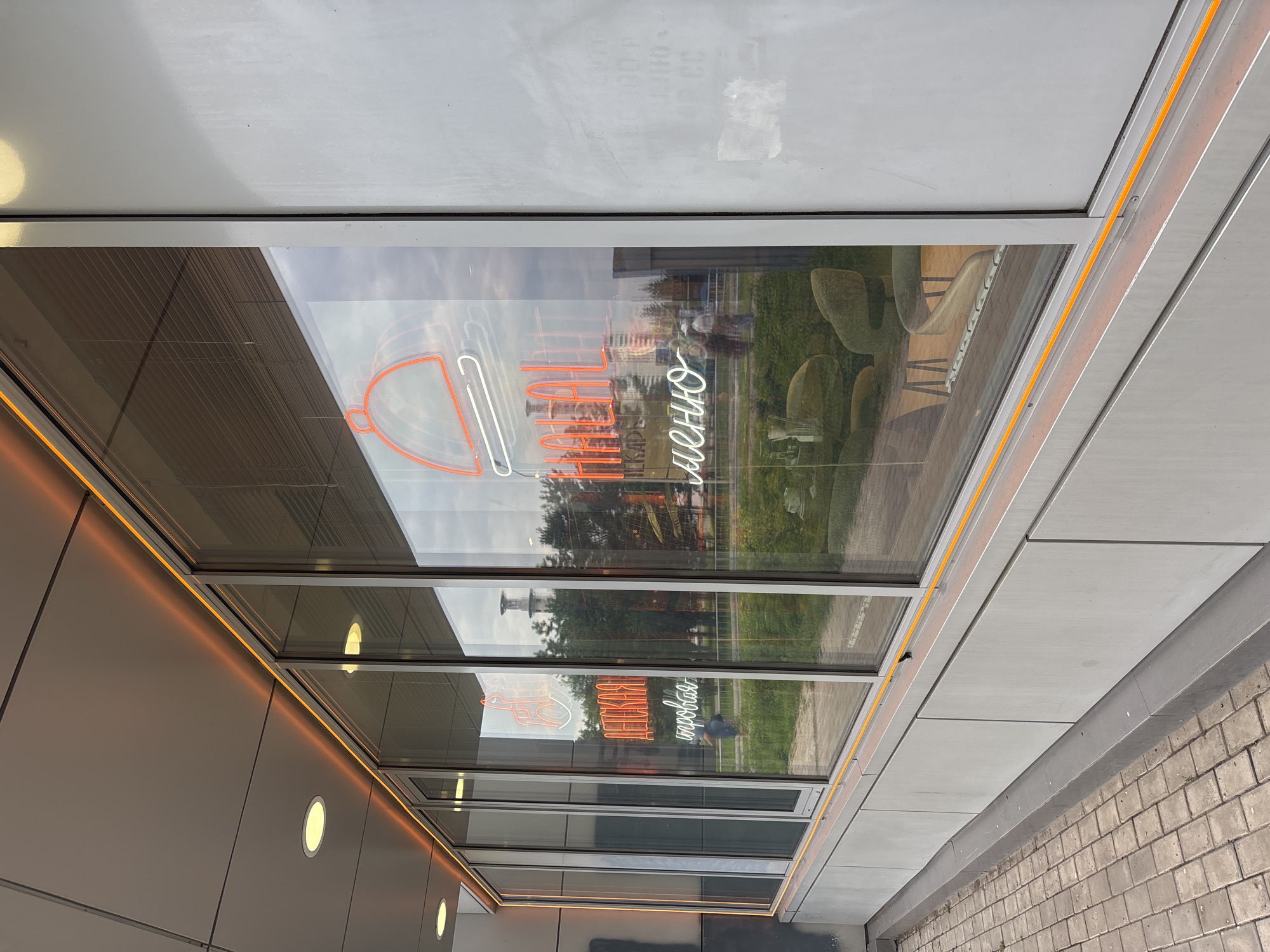 The image shows the exterior of a building with large windows. A neon sign in Russian is visible through the windows. The sign says "ДЕТСКАЯ ИГРОВАЯ" (children's play area) and "HALAL МЕНЮ" (Halal menu). Inside the building, chairs and tables are visible. The exterior of the building is modern, with gray panels and an orange light strip running along the top of the windows. The pavement in front of the building is made of bricks. The sky is overcast. The image is likely taken in Naberezhnye Chelny, Russia, given the Russian text on the sign.