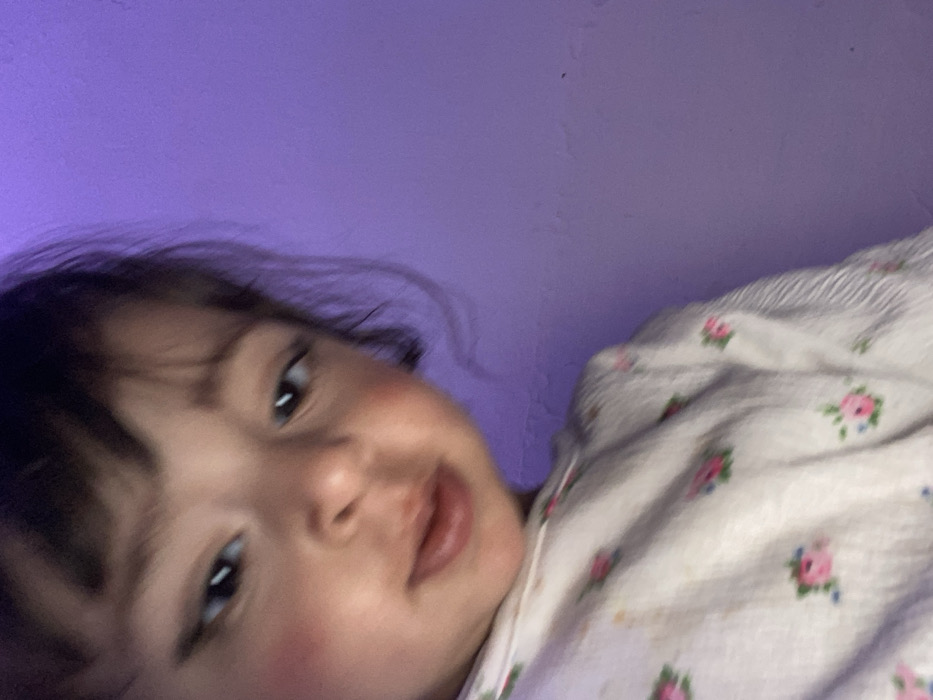 The image presents a close-up, slightly soft-focus depiction of a young child with dark hair, bangs, dark eyes, and pronounced rosy cheeks, looking downwards and to the viewer's left. The child wears a light-colored top patterned with small pink roses and green leaves. The right half of the frame and the background behind the child consist of a solid purple wall, exhibiting minor textural variations or imperfections. The entire scene is bathed in a uniform purple light, suggesting a colored light source. The visual elements within the image do not provide specific contextual information to identify its location as Kandahar, Afghanistan.