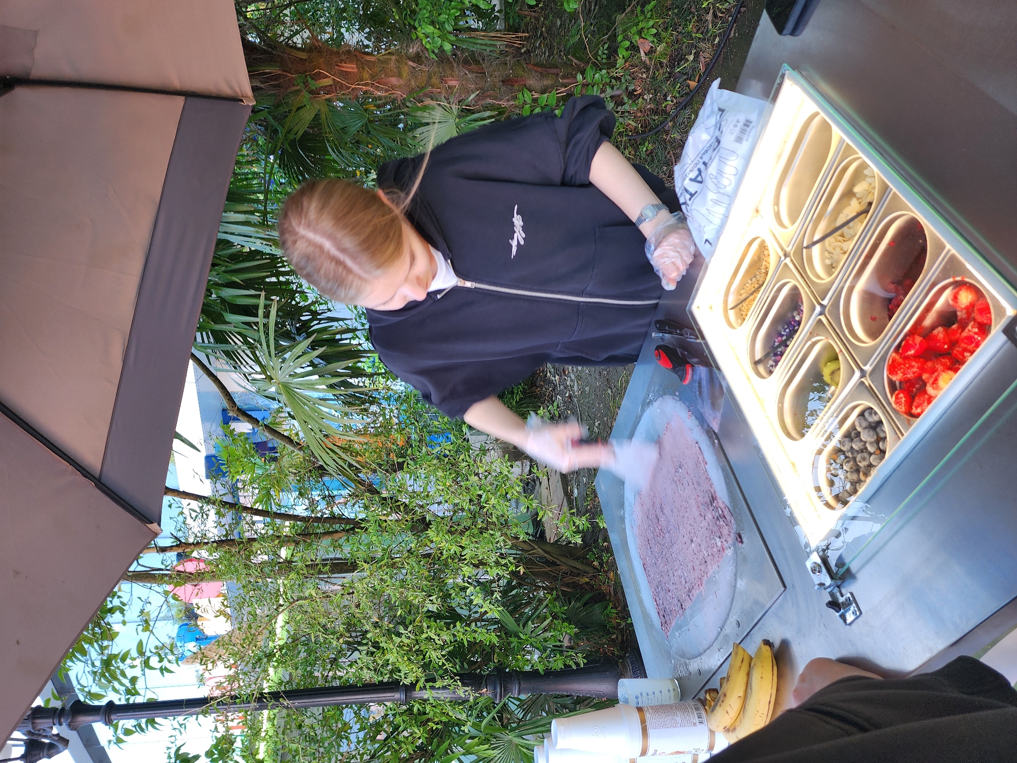 This image captures an outdoor scene at a food stall, likely preparing rolled ice cream, in a location consistent with Sochi, Russia. The perspective is from slightly above and to the left, looking down onto the preparation area.
The main subject is a person with light brown or blonde hair, seen from the back, wearing a black zip-up hoodie featuring a white, stylized script logo on the chest. They have clear plastic gloves on both hands and a silver watch on their left wrist. The person is actively engaged in preparing rolled ice cream on a circular cold plate, using a metal spatula to spread and mix a pinkish-purple substance, likely an ice cream base with fruit.
Adjacent to the cold plate is a lighted display tray holding various toppings for the ice cream. Visible ingredients include a dark brown substance (possibly chocolate), a vanilla bean, sliced almonds, small dark berries (such as blueberries or blackcurrants), kiwi slices, and bright red berries (like strawberries or raspberries). On the counter nearby are two ripe bananas, several white plastic cups, and bottles with some illegible text. A white plastic bag with partially visible Cyrillic text, including "ОЛО" and "АТ", is also present.
The setting is outdoors, sheltered by a large, beige and brown umbrella. The background is filled with lush green foliage, including large palm trees and dense bushes, indicative of a subtropical climate. Glimpses of a bright blue sky and distant buildings with blue and other colored elements are visible through the leaves. A black decorative lamppost stands amidst the greenery. The time of day appears to be daytime, likely bright and sunny, necessitating the use of the umbrella for shade. The atmosphere suggests a relaxed, outdoor culinary experience.