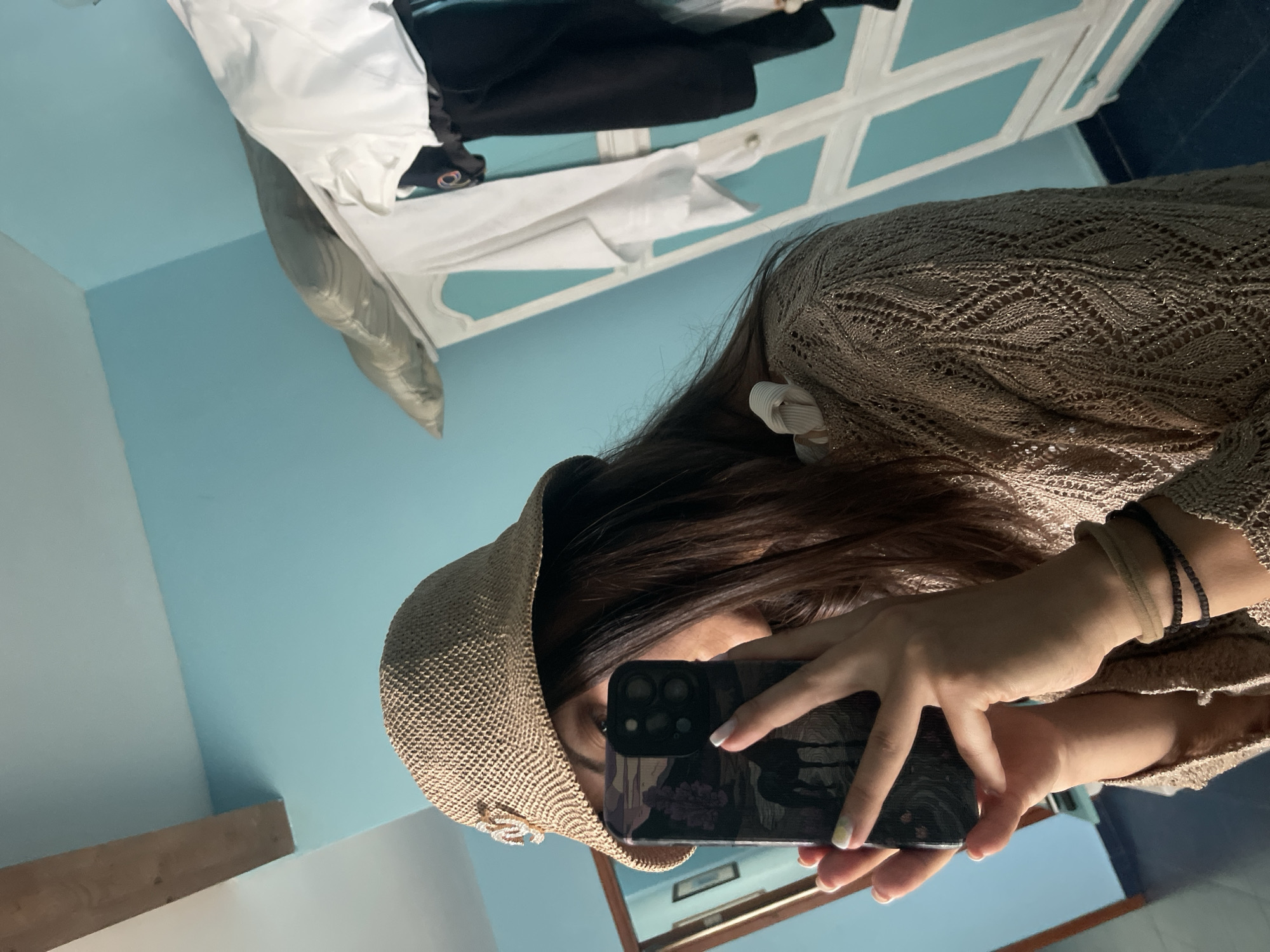 A person is taking a mirror selfie in what appears to be a bedroom. The person is wearing a straw hat and a light brown, open-knit sweater. They are holding a smartphone with a dark phone case that features a nature scene with a horse. The background shows a blue wall, a wooden beam, and a white wardrobe with some clothing hanging on it. There is a mirror on the left side of the image, reflecting another doorway and a picture on the wall. The scene is lit by artificial light, creating shadows.