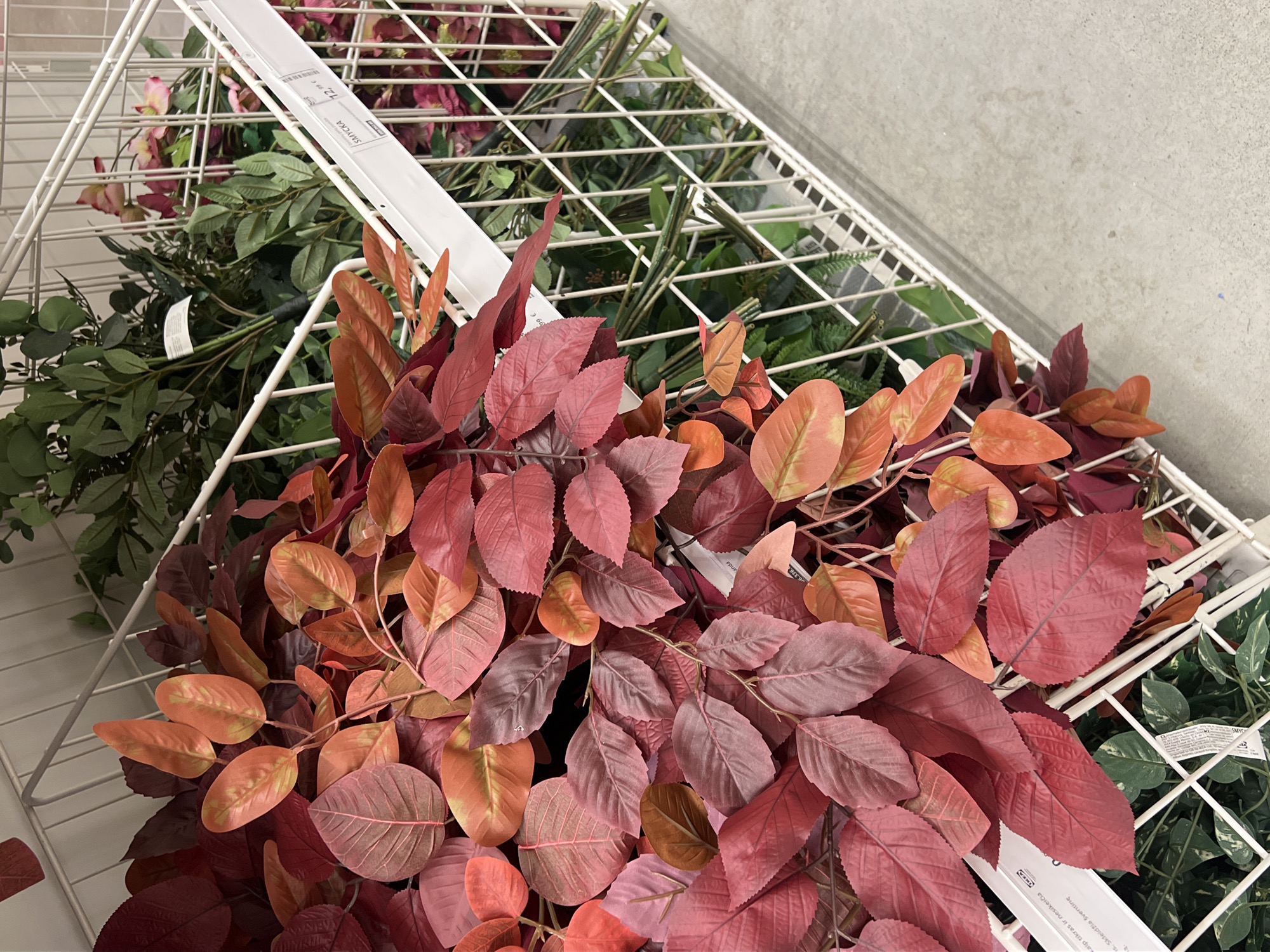 The image shows a close-up view of artificial plants displayed in a white wire rack, likely in a retail store. The foreground is dominated by branches of artificial leaves in vibrant shades of red, maroon, and orange. Some leaves are a deep red, while others transition to a warm orange, creating an autumnal effect. The texture of the leaves is visible, with distinct veins. Behind these, in the middle ground, are bundles of artificial greenery with smaller, darker leaves, and some artificial flowers in shades of pink and darker red. The plants are arranged in a storage or display unit with a white metal frame. Price tags are visible on the shelving, with one prominently displaying "SMYCKA" and "72, 99 €". The background consists of a concrete floor and the continuation of the display shelves. The scene suggests a retail environment for home decor or artificial plants. The presence of the "SMYCKA" product name and "IKEA" logo on a price tag strongly indicates that this image was taken at an IKEA store. While the specific IKEA store location in Vilnius, Lithuania, is not explicitly visible, the context of an IKEA store implies it is within the city limits of Vilnius.