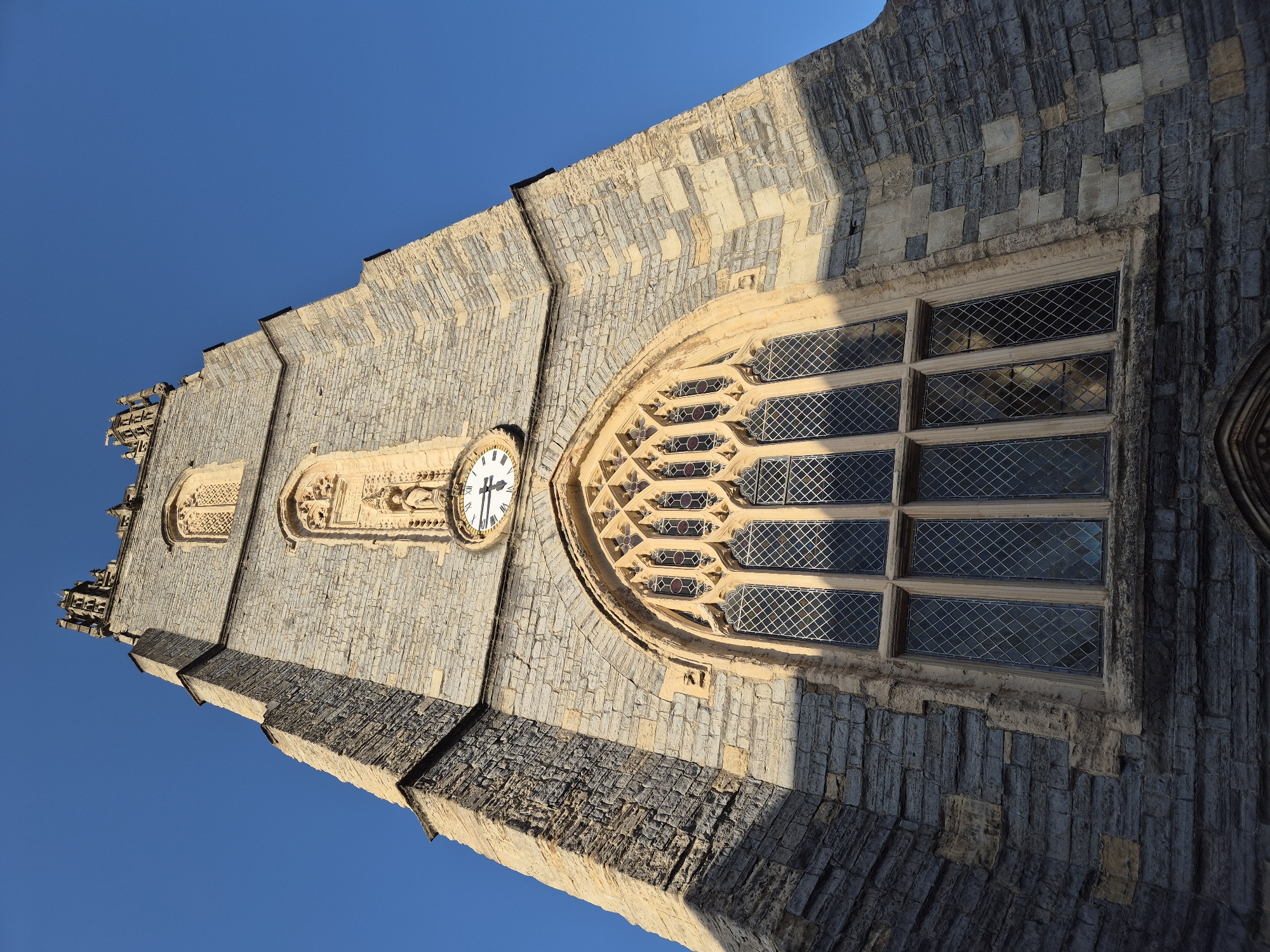 The image shows a low-angle, rotated view of the tower of St. John the Baptist Church in Cardiff, United Kingdom. The tower is constructed of grey stone with lighter-colored accents and contains two windows, the larger one toward the viewer with a detailed, arched frame and diamond-shaped panes. A clock is visible on the tower's facade between the two windows. The sky is clear and blue.