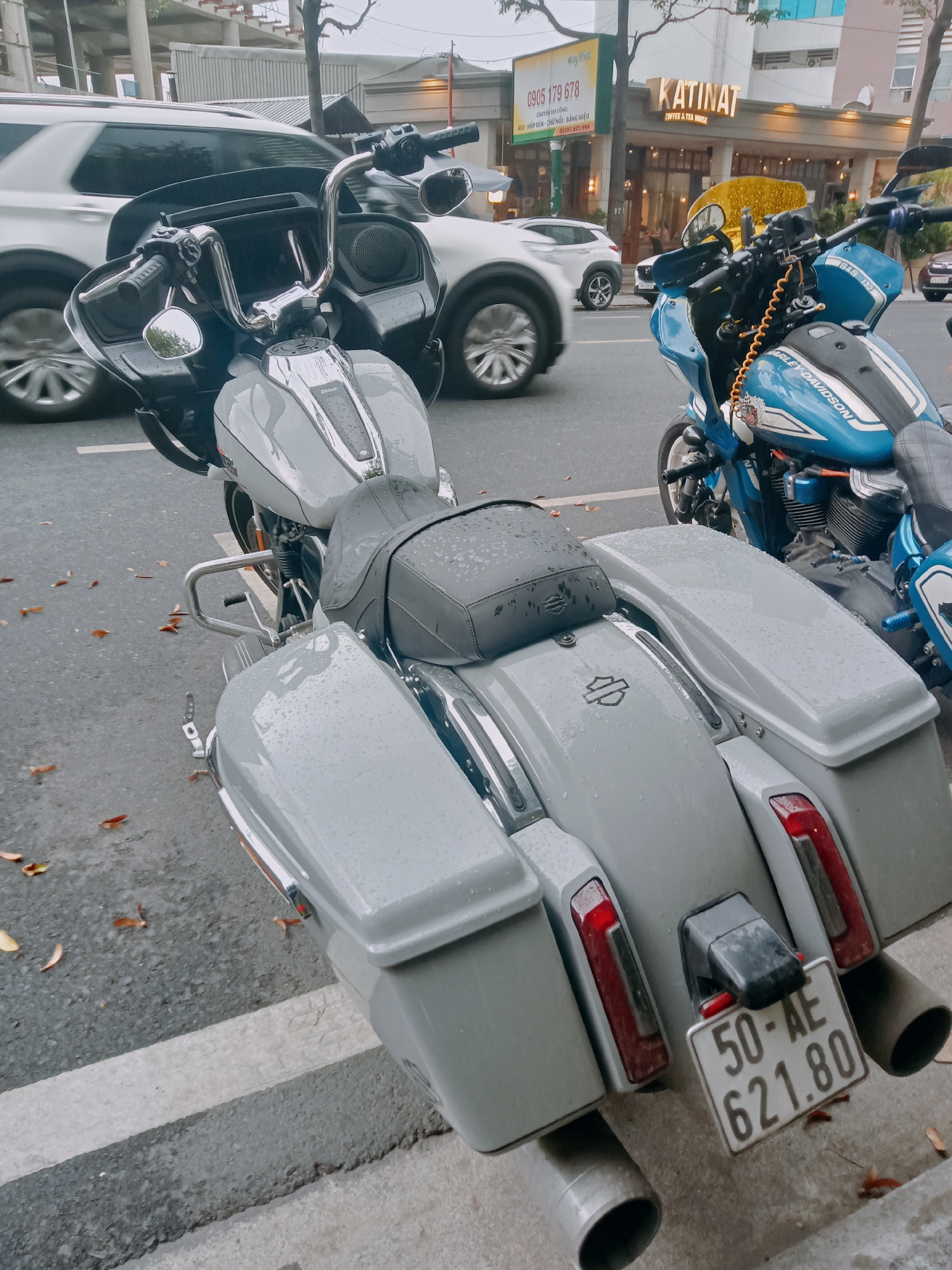 The image shows two motorcycles parked on a city street in Nha Trang Ward, Vietnam. The foreground motorcycle is gray, with saddlebags and a license plate reading "50-AE 621.80". A blue Harley-Davidson motorcycle is parked next to it. In the background, a white car is moving on the street. Above the motorcycles is a sign for "KATINAT COFFEE & TEA HOUSE" and another sign advertising construction services with the phone numbers "0905 179 678" and "02581.872.994". The street is wet, and there are fallen leaves on the ground.
