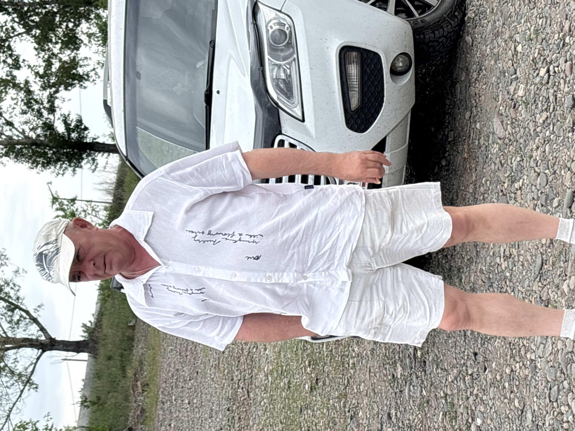 An adult Caucasian male wearing a white cap, a white short-sleeved button-down shirt with text detailing, and white shorts stands on a gravel surface in front of a white SUV. The male has his hands in his pockets and is looking to his left. The SUV has a black bug deflector on the hood and is parked on a gravel road, possibly in an outdoor area. The background consists of trees and vegetation, suggesting a semi-rural or park-like setting. The sky is overcast. The image appears to have been taken in the city of Chernogorsk, Russia, based on the provided context.