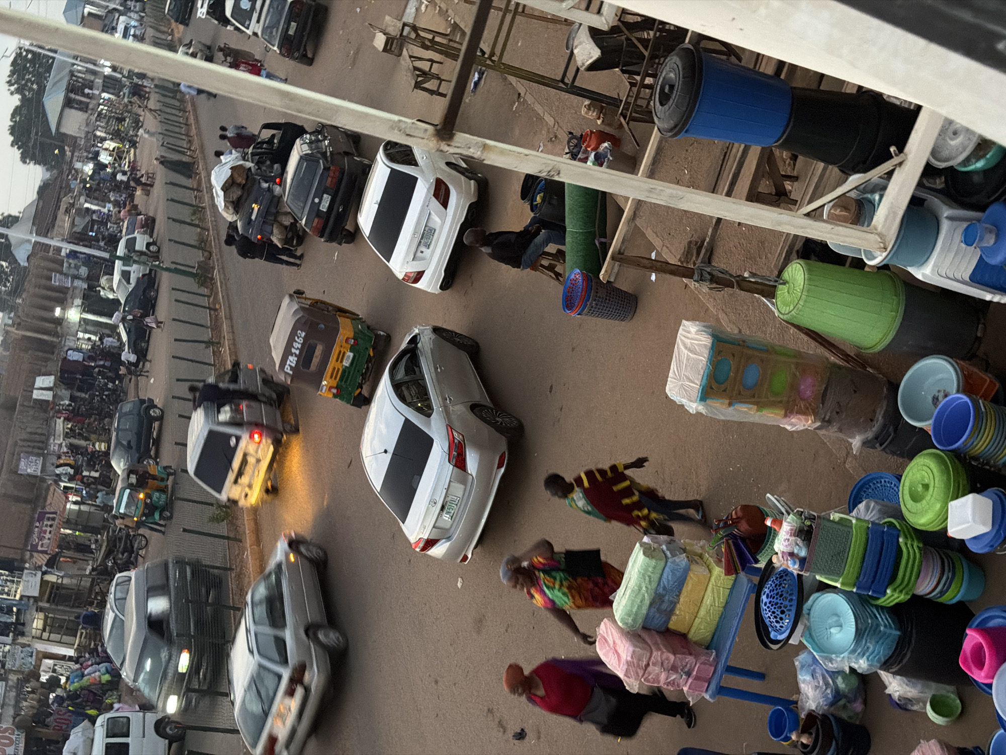 The image captures a street scene in Bukuru, Nigeria, likely a marketplace or busy thoroughfare. In the foreground, various plastic household items are piled on the ground and on a blue plastic table, including stacks of colorful folded fabrics, bowls, buckets, and containers. Three women are walking past the merchandise; one in a red headscarf and purple shawl, another in a vibrant, multi-colored dress, and a third in a dark garment with a striped shawl.
The street is populated with several vehicles, including cars, a minivan, and a three-wheeled auto-rickshaw (Keke Napep). A silver sedan is parked on the right side of the street. A dark colored SUV is visible in the middle distance, as is a white sedan. A man is seated on a stool near some plastic containers on the right side of the frame. Pedestrians are scattered throughout the scene, some walking along the street and others near the market stalls that line the background. The buildings in the background appear to be shops and other commercial establishments. The ground is unpaved in places, with dust visible. The lighting suggests it is either early morning or late afternoon.