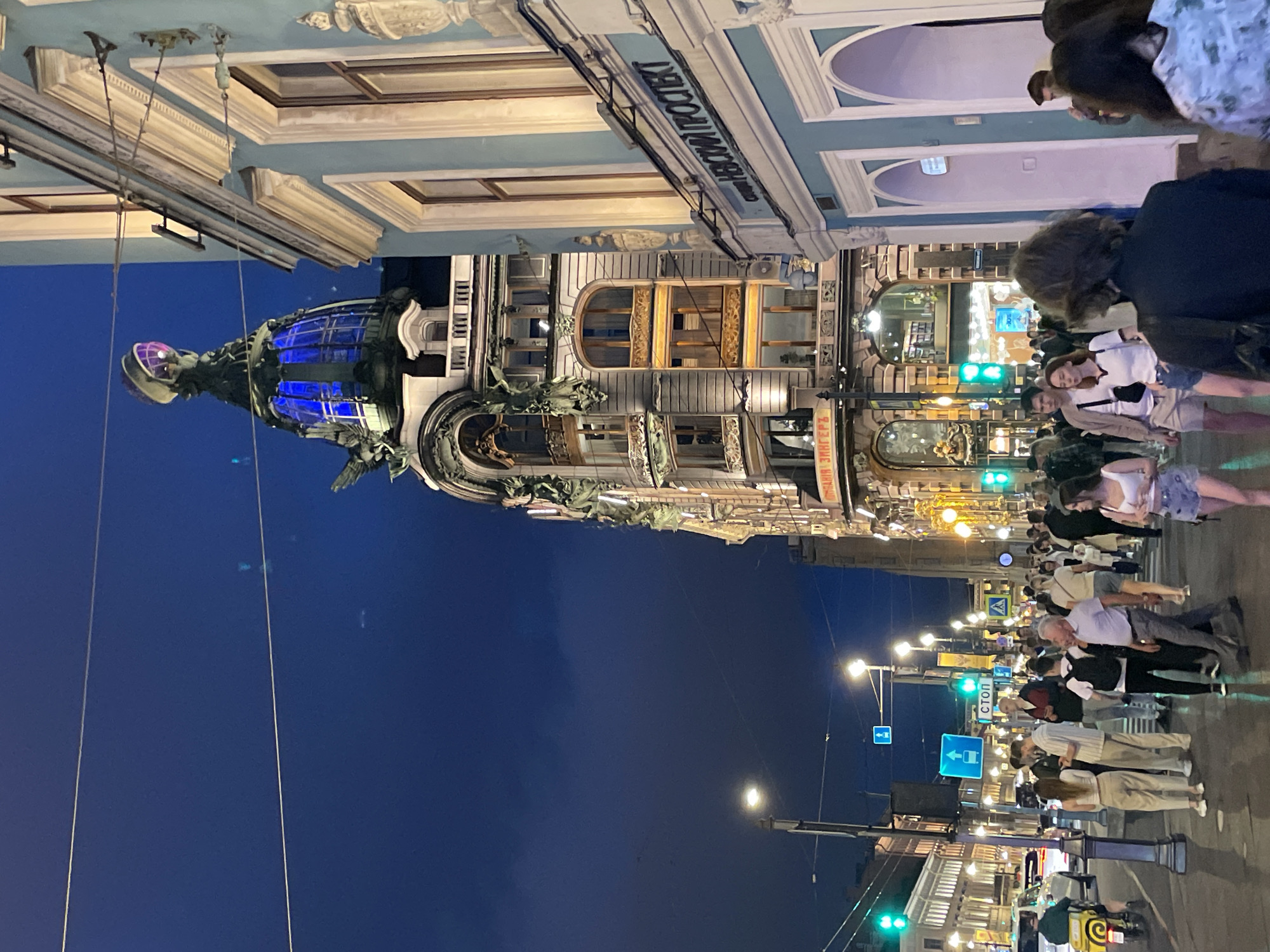 The image depicts a bustling urban street scene at night in Saint Petersburg, Russia. The prominent building on the left is the Singer House (Dom Knigi), an ornate Art Nouveau structure with a distinctive glass dome illuminated in blue light, topped by a purple-lit globe. Adjacent to it on the right is a light blue building with classical architectural elements, featuring a sign in Cyrillic that reads "НЕВСКИЙ ПРОСПЕКТ" (Nevsky Prospekt), definitively identifying the location.
The sky is a deep, dark blue with minimal cloud cover, indicating late evening or night. Numerous streetlights with bright white illumination are active, casting light onto the pavement and street. Several thin overhead wires crisscross the upper sky. Green traffic lights are visible on the left side of the street, and various blue street signs, including a "СТОП" (STOP) sign and directional arrows, are mounted on poles.
The wide pavement is crowded with a diverse group of pedestrians, primarily young to middle-aged adults, dressed in casual modern attire such as t-shirts, shorts, jeans, and light jackets. Individuals are walking in various directions, some singly and others in small groups, with a few appearing to be engaging in conversation or using mobile phones. The pavement surface appears somewhat reflective in places, possibly due to dampness.
On the street to the left, a white car is visible, along with a yellow scooter or moped ridden by a person wearing a green top. Other less distinct vehicles are present further down the street. The Singer House displays intricate architectural details, including statues and illuminated windows, while the adjacent blue building features arched entrances and ornate window frames.