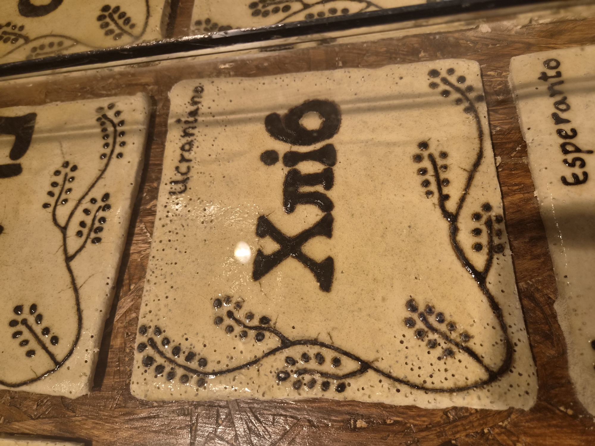 The image shows a close-up of three ceramic tiles with dark lettering and decorative elements, likely displayed on a wooden surface. The tiles appear to be part of a larger installation. The top tile has a decorative pattern of dots and lines resembling branches with berries. The middle tile features a decorative border and the Cyrillic word "Хліб" (Khlib), meaning "bread" in Ukrainian, alongside the word "Ucraniane" written in Latin script. The bottom tile displays the word "Esperanto" in dark lettering. The overall aesthetic suggests a display related to languages or cultural greetings, possibly in a public space. The lighting in the image is warm, and there is a slight sheen on the ceramic surfaces, indicating they might be glazed. The background is a rough wooden texture. The context of Seia, Portugal is not evident from the image itself.