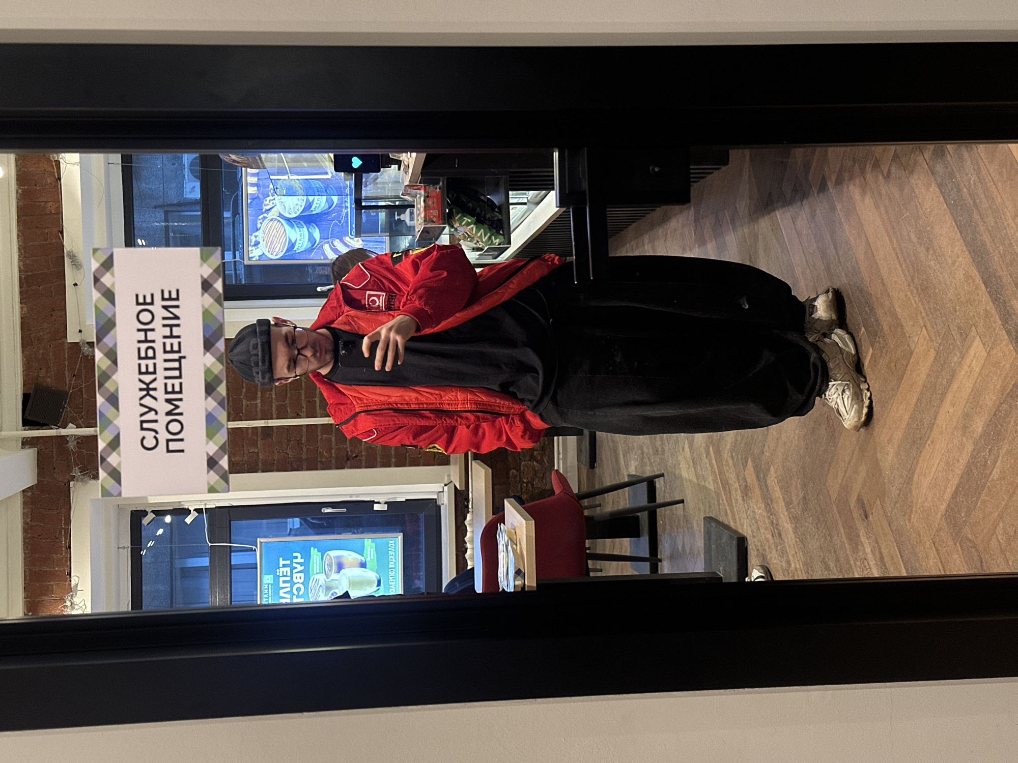 A man stands in a doorway, holding a smartphone. He wears a black beanie, red jacket, and black pants. A sign written in Russian is visible on the glass door; its translation is "Service Premises." The interior of what appears to be a cafe or restaurant can be seen through the door. The floor has a herringbone pattern. Based on the Russian language on the sign and the style of the architecture, the location is likely in Moscow, Russia.