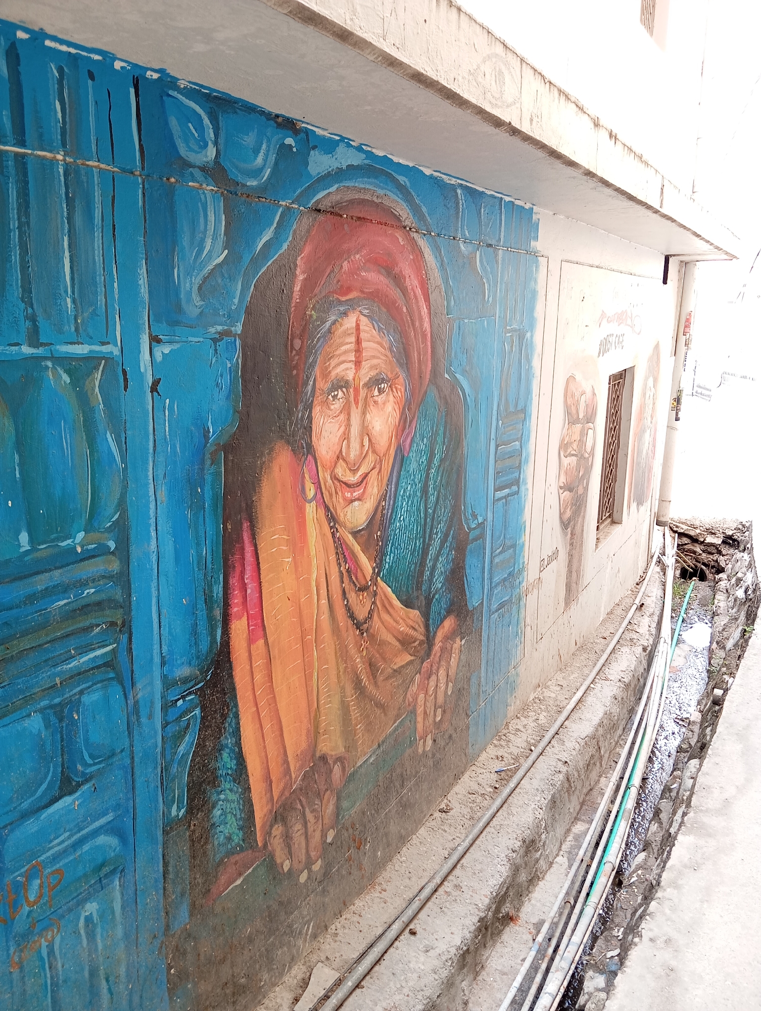 The image shows a mural on an exterior wall in Jonk, India. The main subject of the mural is a painting of an older woman wearing a red headscarf, a sari, and a necklace. She is depicted leaning out of a blue painted window, with her hands resting on the sill. To the right of the woman, there are other painted images on the wall, including a raised fist and a portrait. Below the wall is a narrow, concrete trench with pipes running through it. A drainage ditch runs alongside the wall. The location appears to be a narrow street or alleyway.