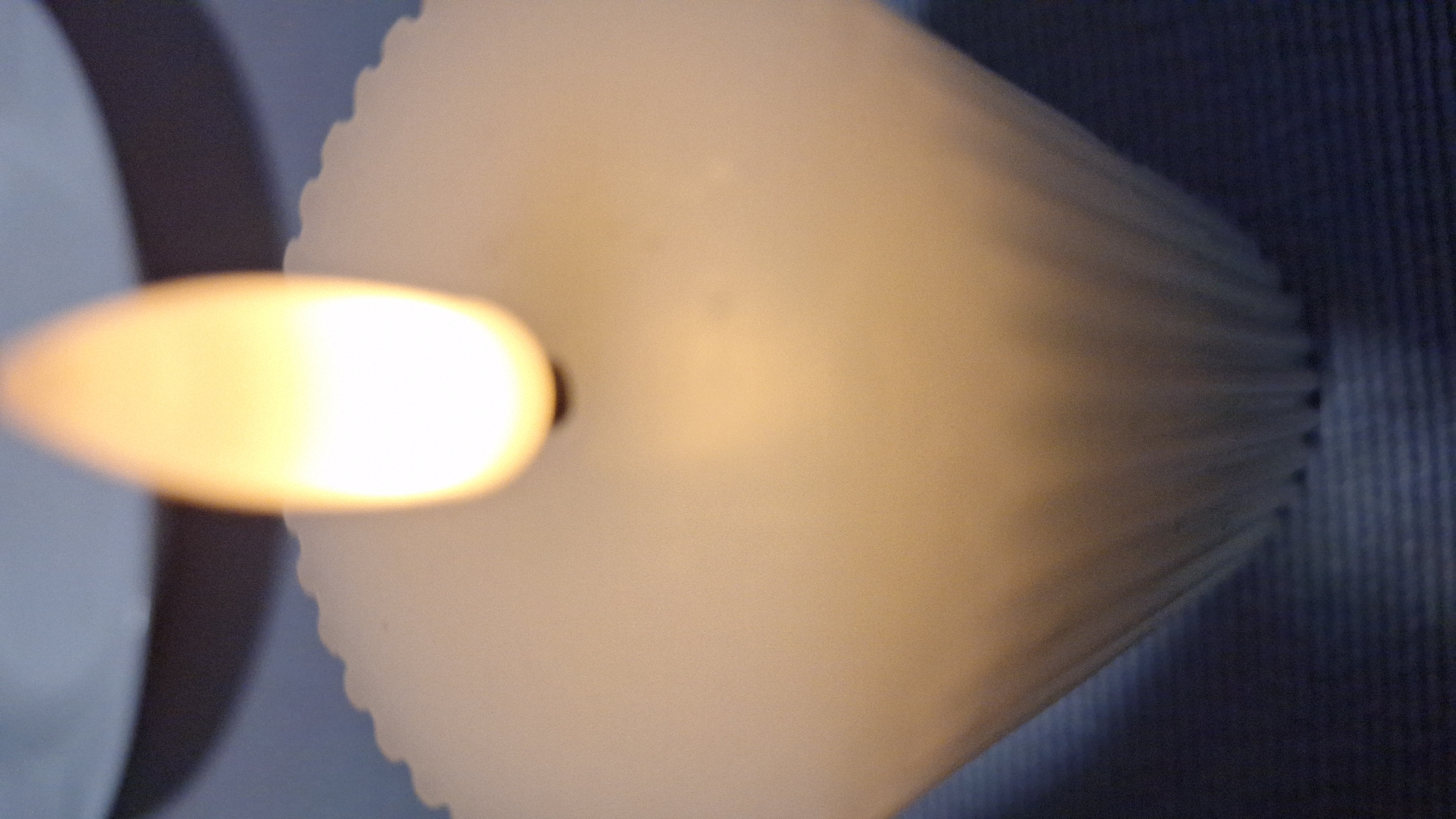 The image depicts a lit candle. The candle is white with a ridged, cylindrical shape. The flame is bright yellow and elongated, positioned at the top of the candle. A partially visible object, possibly another candle or container, is on the left side of the frame. The background appears to be a dark, textured surface, likely a wall. There is no information in the image to indicate the location in Blagoevgrad, Bulgaria.