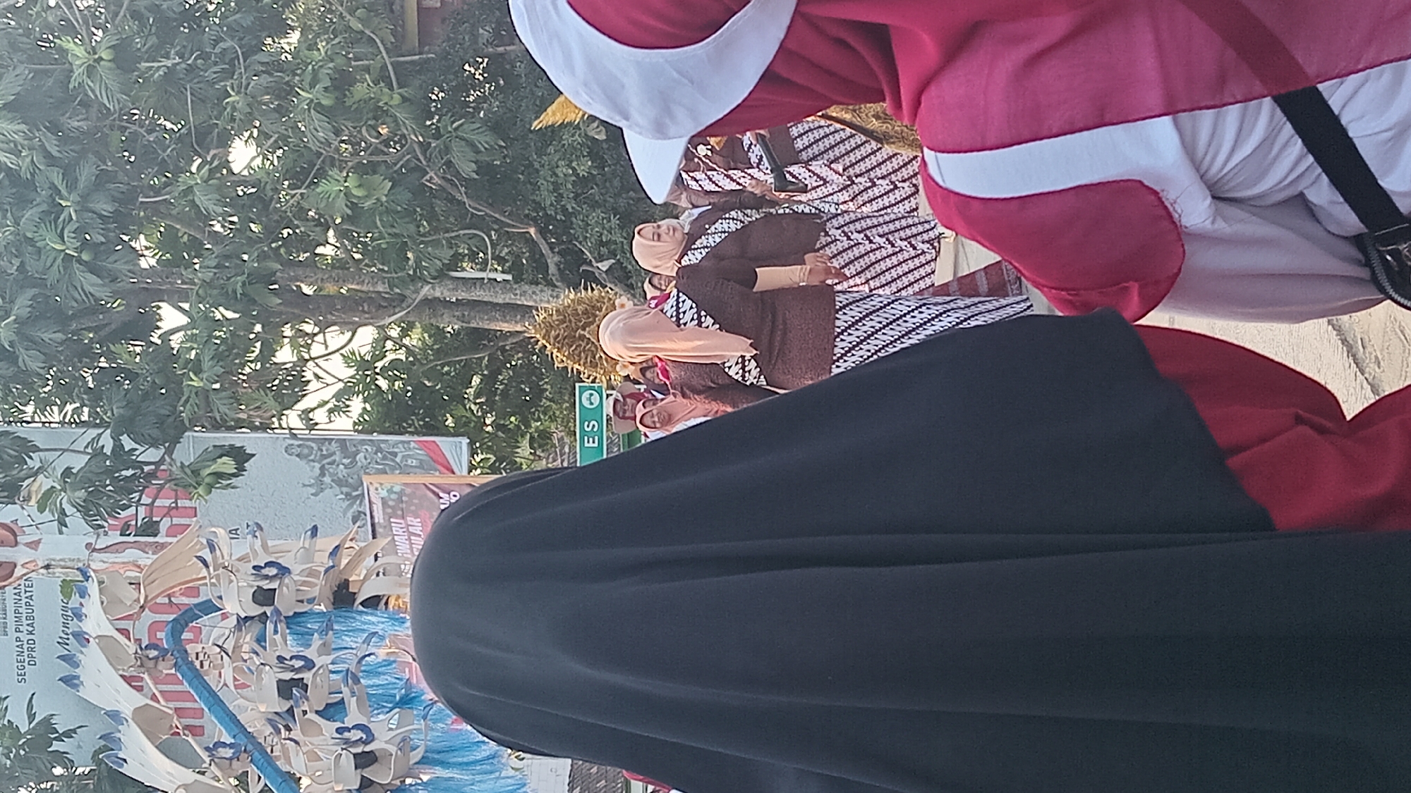 The image depicts a street scene in Kedungwaru, Indonesia, likely during a cultural event or parade. In the foreground, two individuals are partially visible from behind. The person on the left wears a black hijab and a red garment underneath. The person on the right is wearing a red hijab with white trim and a white garment.
In the middle ground, several women are dressed in traditional attire. They wear light brown tops with intricate black and white patterned skirts. Some of them have beige headscarves. They appear to be walking or standing in a procession.
In the background, a large banner with text in Indonesian is visible, featuring the words "SEGENAP PIMPINAN DPRD KABUPATEN" and "Mengue." Another banner or display features artwork or graphics related to a cultural theme. Trees with dense foliage, some bearing fruit, are also present. The ground appears to be paved. The overall lighting suggests it is daytime, possibly late afternoon, given the warm tones and shadows. A sign with the letters "ES" is also visible.