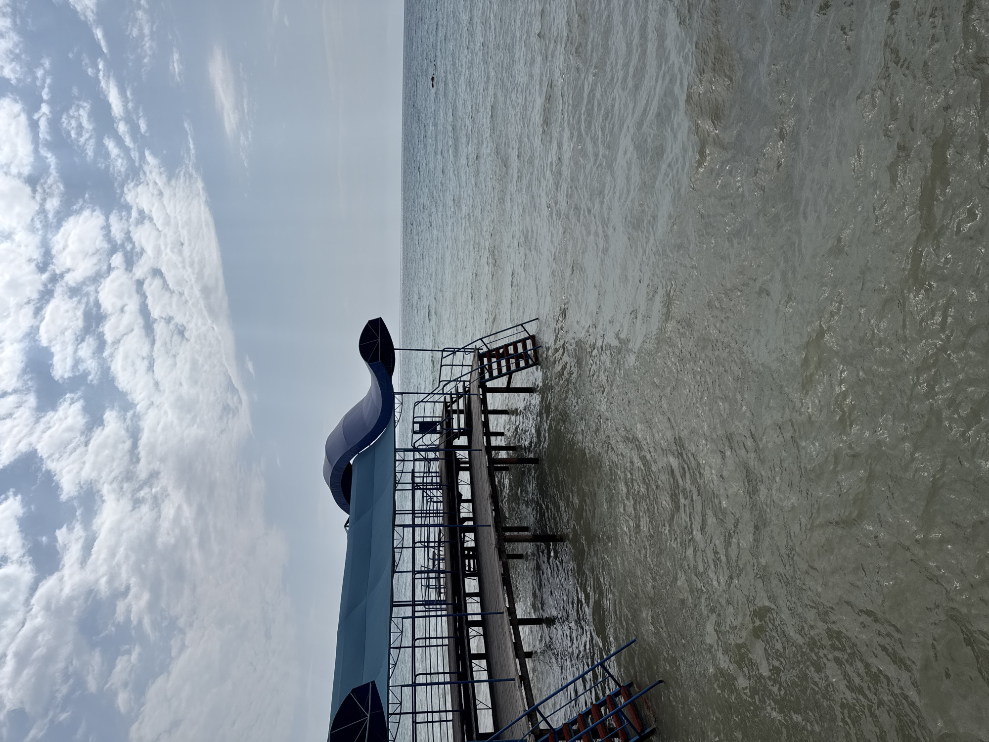 This is a medium shot, eye-level, outdoor, natural light, wide-angle photograph of a wooden pier extending into a body of water. The pier is covered by a blue awning supported by metal poles. There are blue metal railings along the sides of the pier, and wooden steps leading down to the water on both the left and right sides. The water is choppy and appears murky. In the distance, the horizon is visible, with a pale blue sky filled with white, scattered clouds. The scene appears to be in the city of Sary-Oy, Kyrgyzstan, likely a coastal or lakeside area, possibly a resort or public access point. There are no people visible in the photograph.