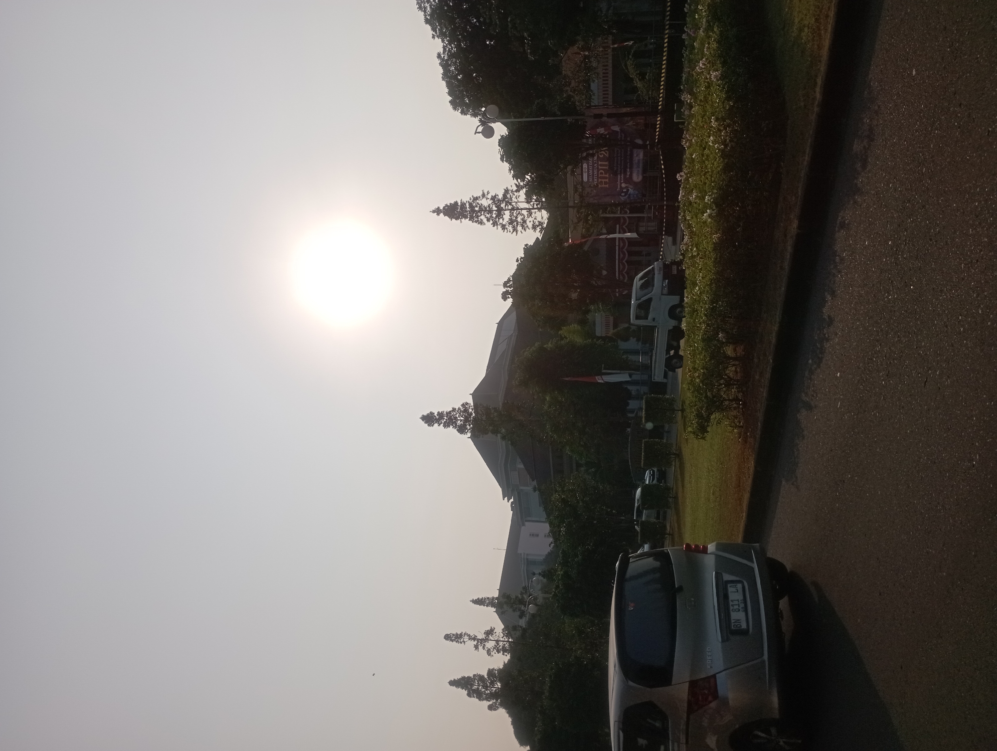 The image depicts a sun-drenched outdoor scene in Depok, Indonesia, likely near a government building or educational institution, given the architecture. The sun is prominently positioned in the upper center of the frame, casting a bright glare that washes out the sky. In the foreground, a silver minivan is parked on an asphalt road, facing left, with its rear visible. The license plate reads "BN 831 LA". To the right of the minivan is a neatly manicured lawn with low hedges and a few scattered flowering plants. Beyond the lawn, a small white flatbed truck is parked. Further in the background, a large building with a dark roof and several floors is visible, partially obscured by lush green trees. One distinctive tall, slender evergreen tree stands out in the middle ground. There are also several flags and banners visible, including one with text that appears to be in Indonesian, suggesting an event is taking place. The overall atmosphere is bright and somewhat hazy, characteristic of a sunny day with perhaps some atmospheric dust or pollution.