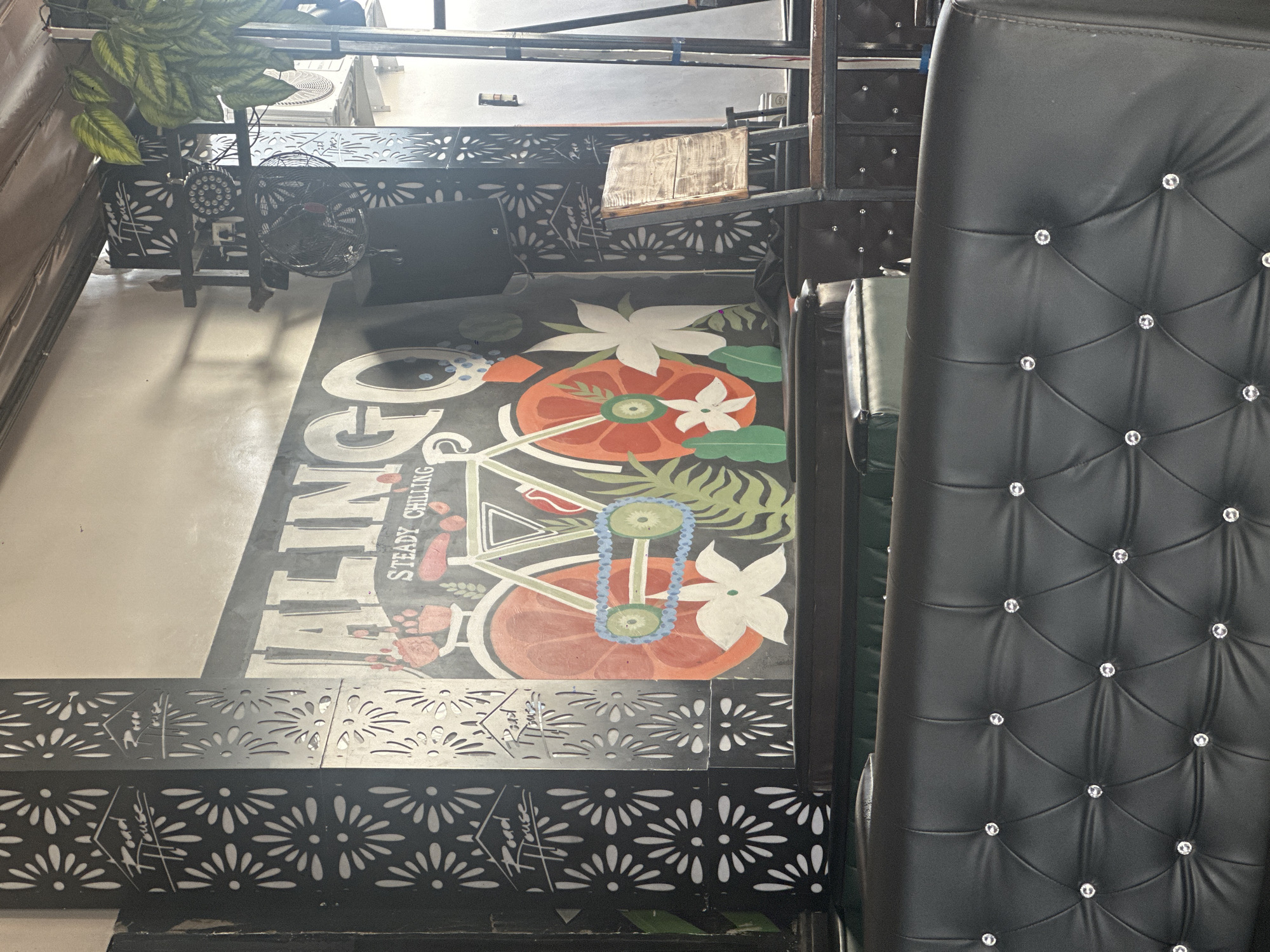 The image depicts an indoor scene, likely a restaurant or cafe, with a clear focus on the floor artwork and seating. The floor features a large, stylized graphic with bold lettering that spells out "GOING STEADY CHILLING". Interspersed with the text are vibrant illustrations of a sliced orange, white flowers, green leaves, and a lei necklace. The artwork appears to be painted directly onto the floor, creating a colorful and engaging design.
Adjacent to the floor graphic, to the right of the frame, is a black upholstered chair with a tufted back, adorned with numerous sparkling embellishments that resemble diamonds or crystals. The chair is positioned to suggest a seating area within the establishment.
In the background, decorative metal screens with a floral pattern are visible, acting as dividers or wall embellishments. Above these screens, industrial elements like air conditioning units and exposed ceiling structures are present, indicating a practical and possibly functional design for the space. A metal railing runs across the top of the image. A small fan is visible in the upper left quadrant, suggesting an effort to circulate air in the space. A potted plant with green leaves adds a touch of greenery to the upper left corner.
There is no clear indication of the time of day or weather. The setting appears to be indoors, and the overall impression is one of a casual and potentially lively establishment, with the artwork and chair design contributing to a unique aesthetic. The visible text is "GOING STEADY CHILLING." The location context is Howayi, Nigeria.