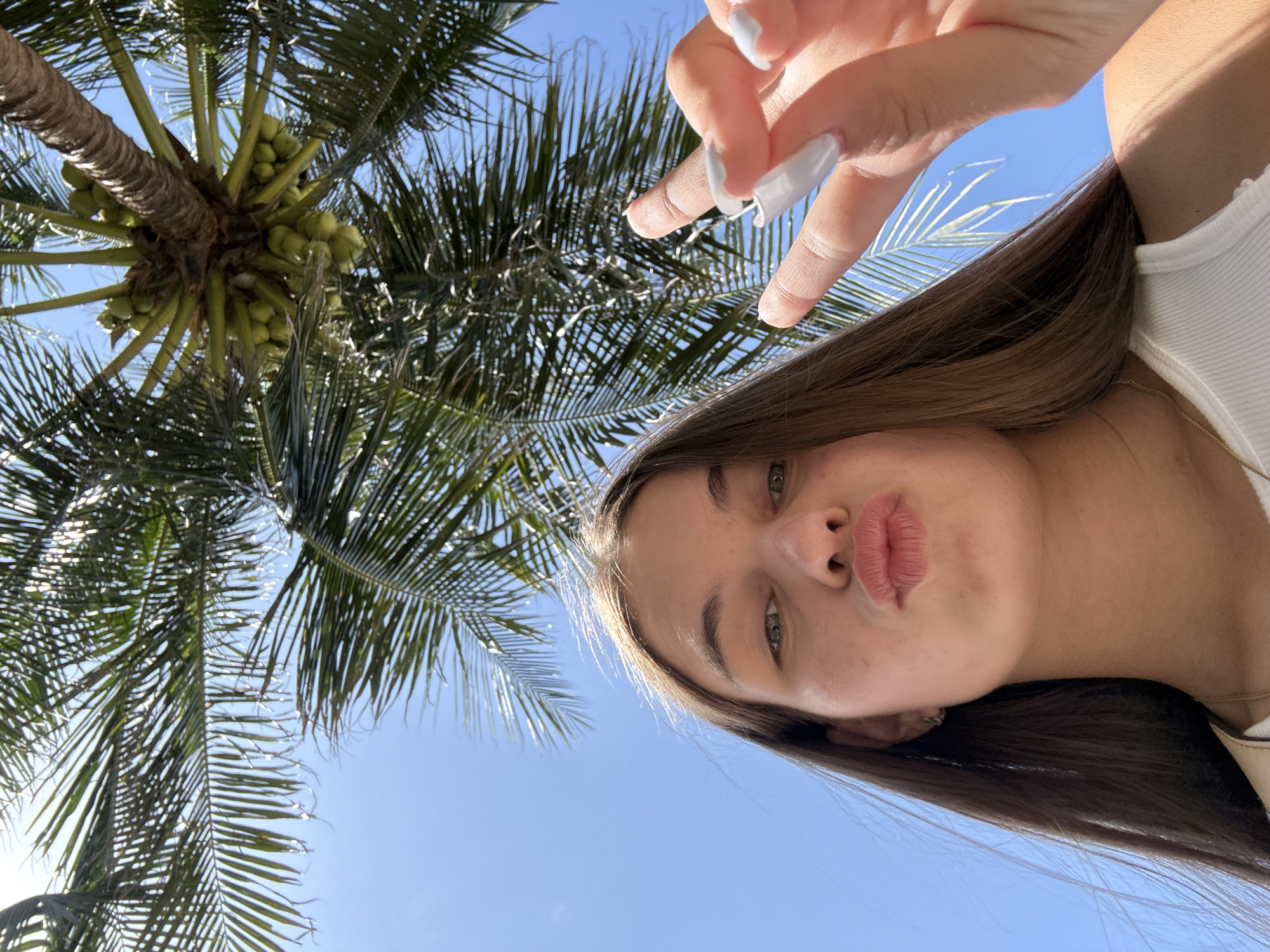 The image shows a young woman with long brown hair, taken from a low angle, with her face turned towards the camera. She is making a kissing face and holding up two fingers in a peace sign with her right hand, which has light blue nail polish. She is wearing a white tank top and a gold necklace. Above her, the fronds of a palm tree are visible, with green coconuts clustered on the trunk. The sky is a clear blue. The image does not contain any information that can indicate the location within Nha Trang, Vietnam.