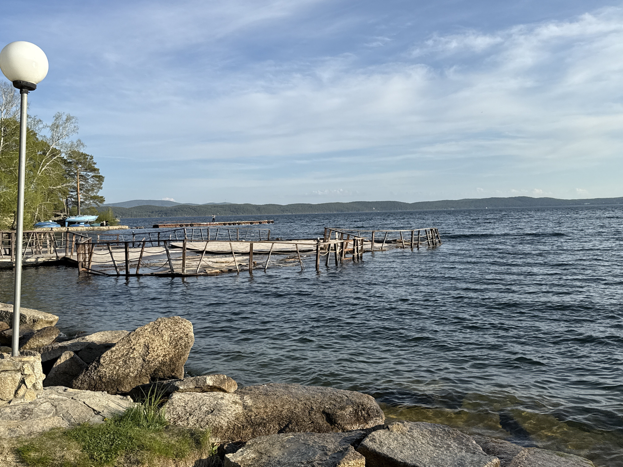 The image depicts a waterfront scene at Zolotoi Plyazh, Russia. A pier or dock extends into a large body of water, likely a lake or reservoir, with low-lying hills or mountains visible in the background. The pier's construction is partially damaged or incomplete, with missing sections of its wooden railing.
In the foreground, large rocks and boulders line the shoreline, with some sparse green vegetation growing among them. A lamp post with a spherical light fixture is positioned on the left side of the frame, near the rocky shore. Further along the pier, a person is standing. On the left is a light-blue boat. The sky is lightly clouded.