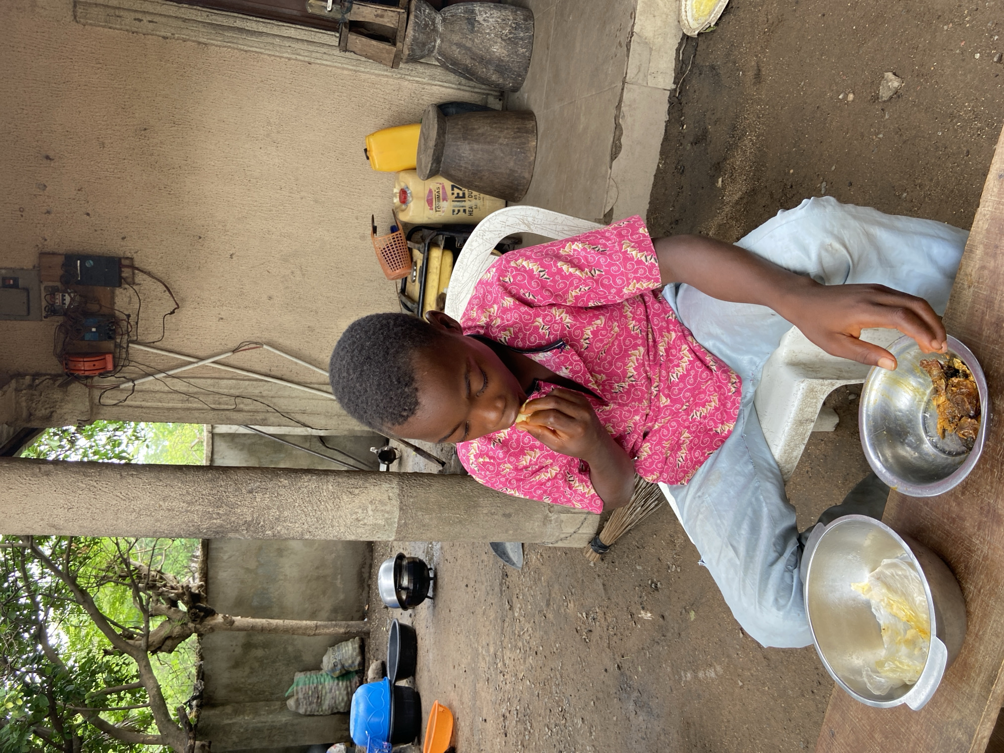 The image captures a young person of African descent eating outdoors in what appears to be Mararaba, Nigeria. The individual, wearing a pink patterned shirt and light blue trousers, is seated on a white plastic chair at a wooden table. In front of them are two metal bowls; one contains food being eaten and the other contains food wrapped in plastic. The location is a yard with a dirt ground and partial shelter created by supports and a roof. Visible in the background are various household items and food prep items, including pots, buckets, and wooden tools, alongside electrical wiring and a concrete wall. Trees and natural vegetation are also visible.