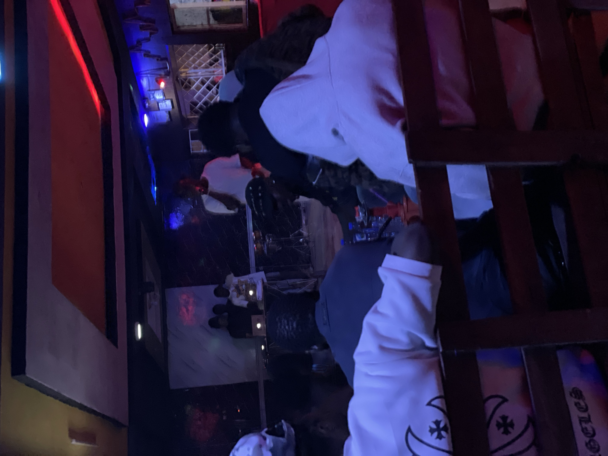 The image depicts an indoor scene, likely a club or lounge, illuminated by a mix of red, blue, and white lights. In the background, three individuals are standing near a table with a laptop, possibly engaged in a DJ set or performance. One person is holding a microphone. In the foreground, several people are seated, facing away from the camera, and appear to be watching the performers. The overall ambiance suggests a social gathering. The location is identified as Durimi, Nigeria.