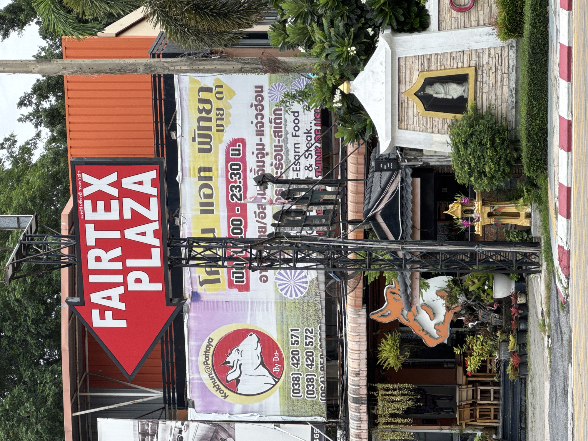 The image shows a street scene in Pattaya City, Thailand, featuring a prominent red arrow sign pointing left, with white text reading "FAIRTEX PLAZA". Below this sign, a large banner advertises "Kokhun@Pattaya" with a cow's head logo and phone numbers, as well as "เอก พัทยา บาย ดา" in Thai script. The banner also lists operating hours from 11:00 to 23:30. Further down, there is a building with a tiled roof and decorative elements, including a statue in a niche and surrounding greenery. Several potted plants are arranged along the sidewalk, and a yellow and red striped curb is visible. The overall environment suggests a commercial area, likely a street with shops and restaurants.