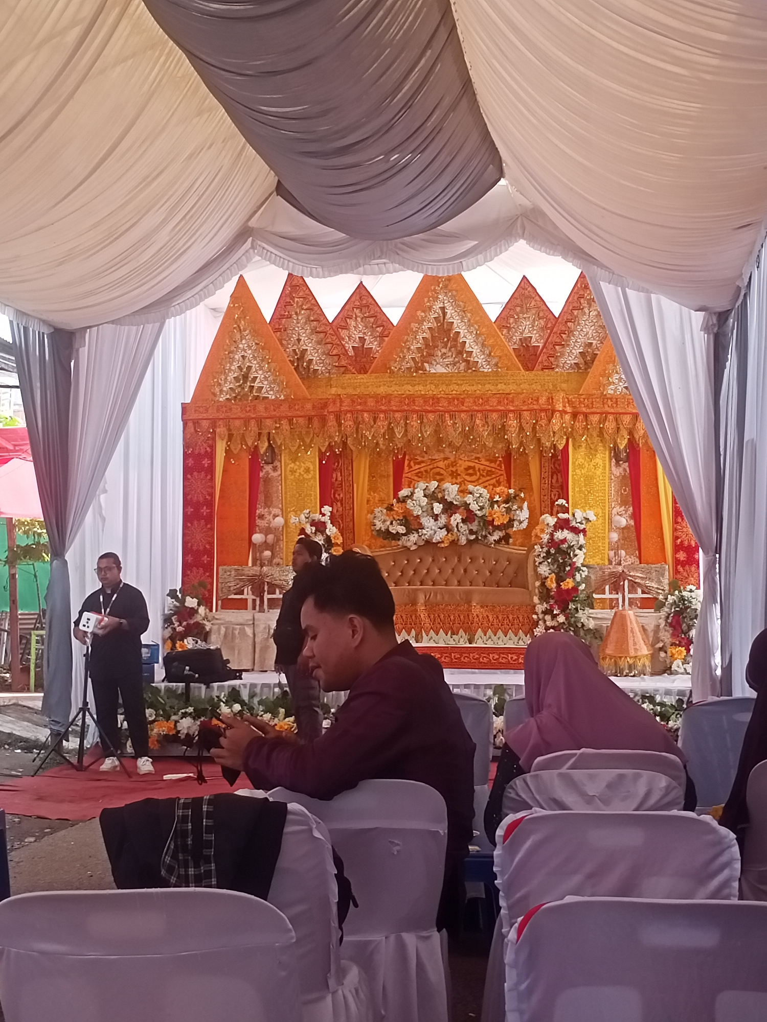 The image shows a gathering under a draped tent structure, likely a wedding or similar event, in Keudematangkuli, Indonesia. The tent is decorated with white and grey fabric draping from the ceiling, and features an ornate, orange and gold backdrop, likely the dais for the event's principals. In front of this backdrop is a decorated couch and various floral arrangements.
Several people are visible. On the left, a man stands holding a device, possibly a microphone or remote control, and seems to be addressing the audience. In the foreground, several seated individuals are visible from the shoulders up, some facing forward and some turned to the side. A woman wearing a pink headscarf is also seated amongst them.
The environment suggests an outdoor location, as evidenced by the glimpse of trees and buildings in the background. The event is staged on a red carpet, and the audience is seated in rows of white chairs.
