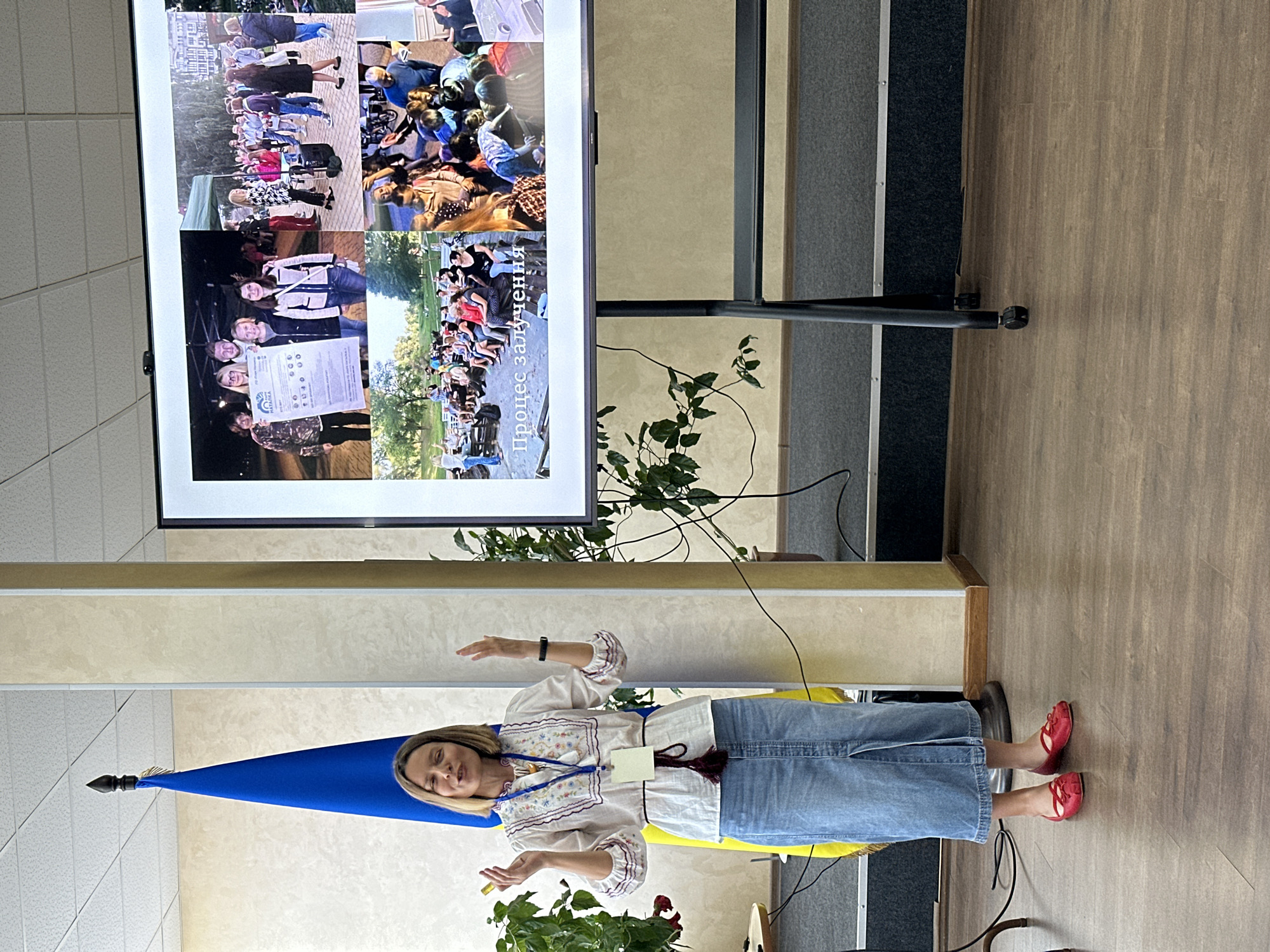 A woman stands in front of a large screen displaying a collage of photos. The screen shows various images of people engaged in different activities. The woman is wearing a traditional embroidered shirt, jeans, and red shoes. She is gesturing with her hands as if speaking or presenting. Behind her, a Ukrainian flag is visible. The scene appears to be indoors, possibly a conference room or auditorium, with wooden flooring and beige walls. Plants are present in the foreground. The overall mood seems to be one of presentation or education. The visible text on the screen reads "Процес залучення". The location is Myronivka, Ukraine.