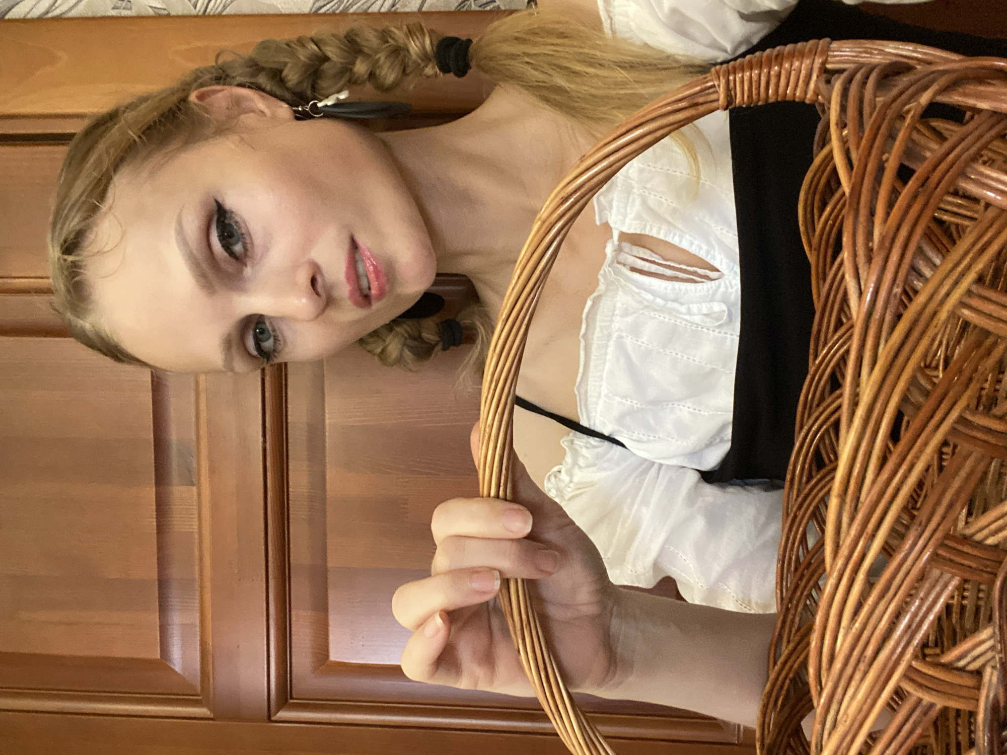A young woman with blonde hair in two braids, wearing a white off-the-shoulder top and a black dress, holds a wicker basket. She is looking at the camera, her head tilted slightly. Her makeup includes eyeliner and lipstick. She is indoors, in front of a wooden door. The scene appears to be in Lida, Belarus, though no specific landmark is visible.