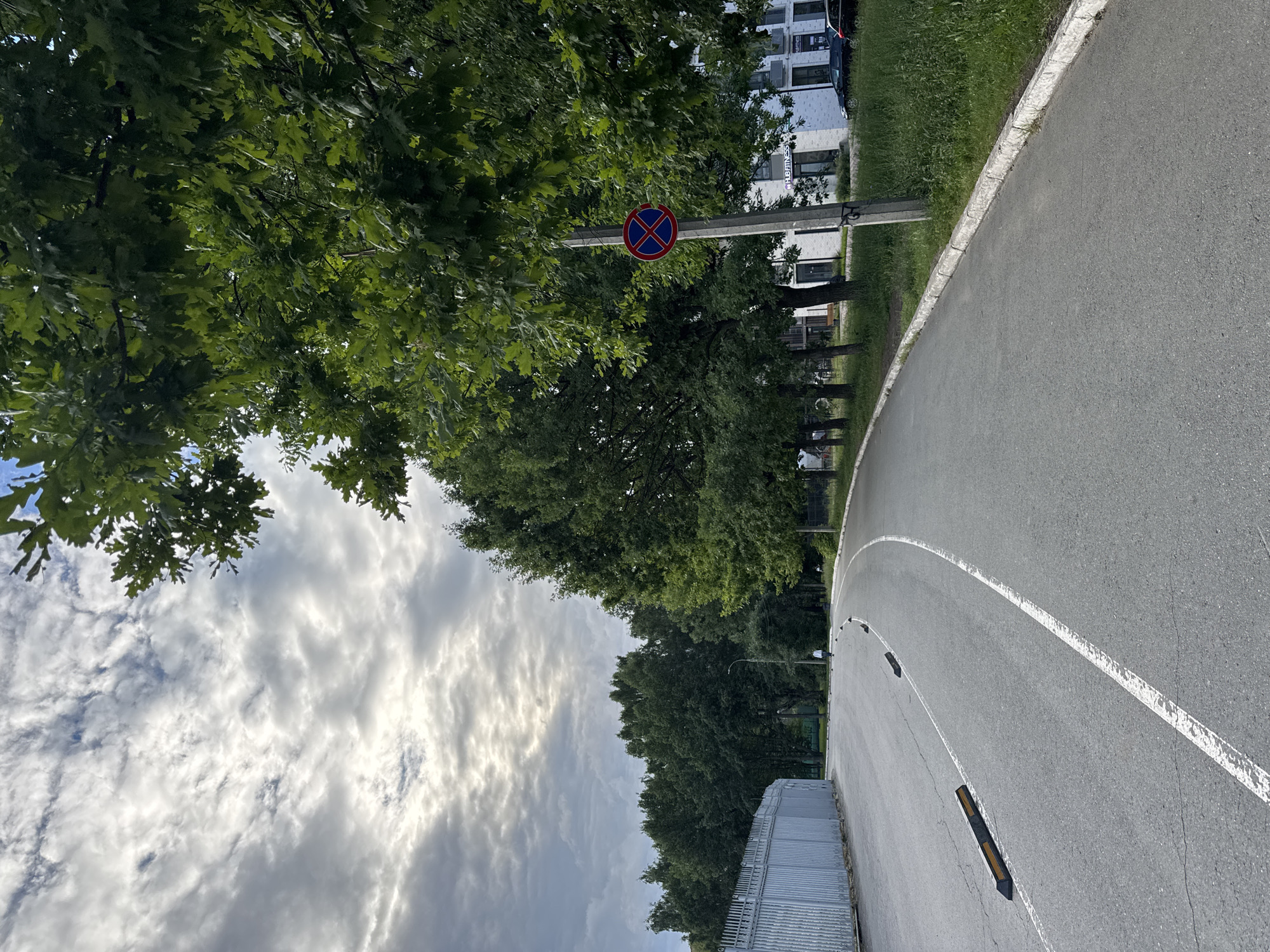 The image shows an asphalt road with white markings, curving to the right. Along the left side of the road is a tall, metal fence. On the right side of the road, there is a concrete pole with a circular sign depicting a red border and a blue interior with a red "X" symbol, indicating a no parking zone. Adjacent to the road is a strip of green grass, with a line of trees behind it. In the background, there are several modern buildings, one with the text "HUB FITNESS" visible. Several cars are parked in front of the buildings. A person is walking on the road in the middle distance. The sky is overcast with scattered clouds. The scene appears to be an outdoor public space.