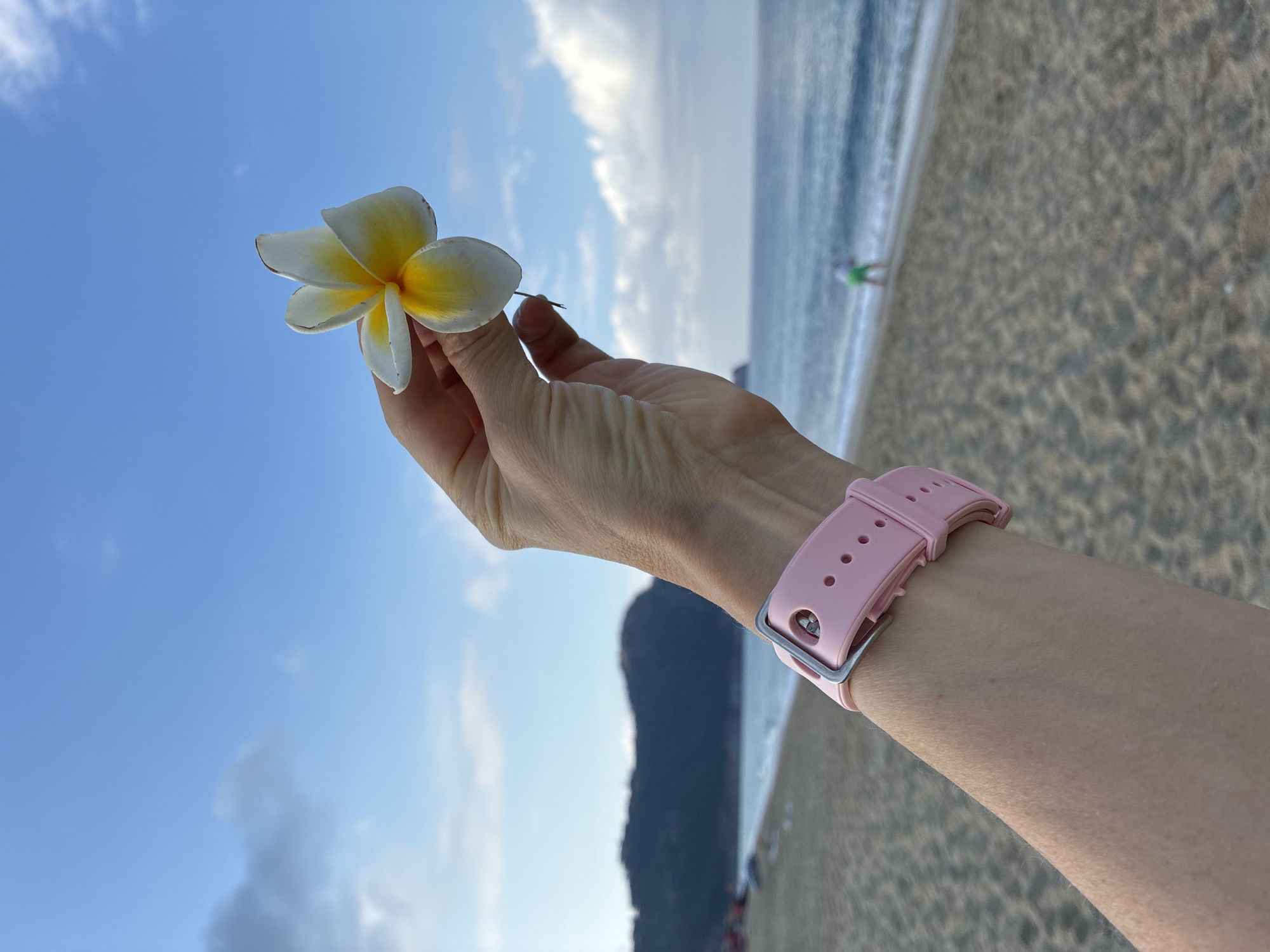 A person's left arm, adorned with a pink smartwatch, is in the foreground of the image. The arm is holding a single white and yellow plumeria flower up towards a bright blue sky with scattered clouds. In the background, a sandy beach stretches towards the ocean, with gentle waves lapping the shore. A distant, dark, mountainous landscape forms the horizon. The scene appears to be a serene beach environment, potentially in Alanya, Turkey, given the visual cues of a coastal city.