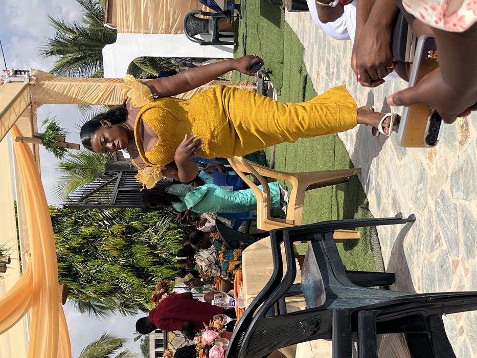 The image captures an outdoor social event under bright daylight in Kumasi, Ghana. The scene features multiple individuals gathered beneath light orange fabric canopies adorned with green foliage.
In the mid-ground, a woman with dark skin, styled dark hair, wears a vibrant mustard yellow traditional-style dress. The dress features a beaded off-shoulder sweetheart neckline bodice with ruffled sheer yellow fabric detailing on the shoulders. She is walking from left to right across a ground composed of irregular flagstones and artificial green turf, holding a dark-colored smartphone in her left hand. She wears white flat sandals and appears to be looking towards the right side of the frame.
To the left, several people are visible. A man in a dark red long-sleeved shirt stands near a round table covered with a beige tablecloth. The table displays a prominent pink floral centerpiece, empty glasses, and a plastic water bottle. Multiple individuals, some in traditional Ghanaian fabrics, are seated at this table and in the background. A woman in a light blue traditional dress is seen standing further back, holding a smartphone.
The background reveals a large, dark metal gate, dense green foliage, and palm trees under a clear sky. Various plastic chairs in dark blue/green, beige, and black are distributed throughout the event space, some occupied and some empty. In the immediate foreground, the hands of two individuals are partially visible, holding smartphones, one of which has a clear case and a triple camera lens array.