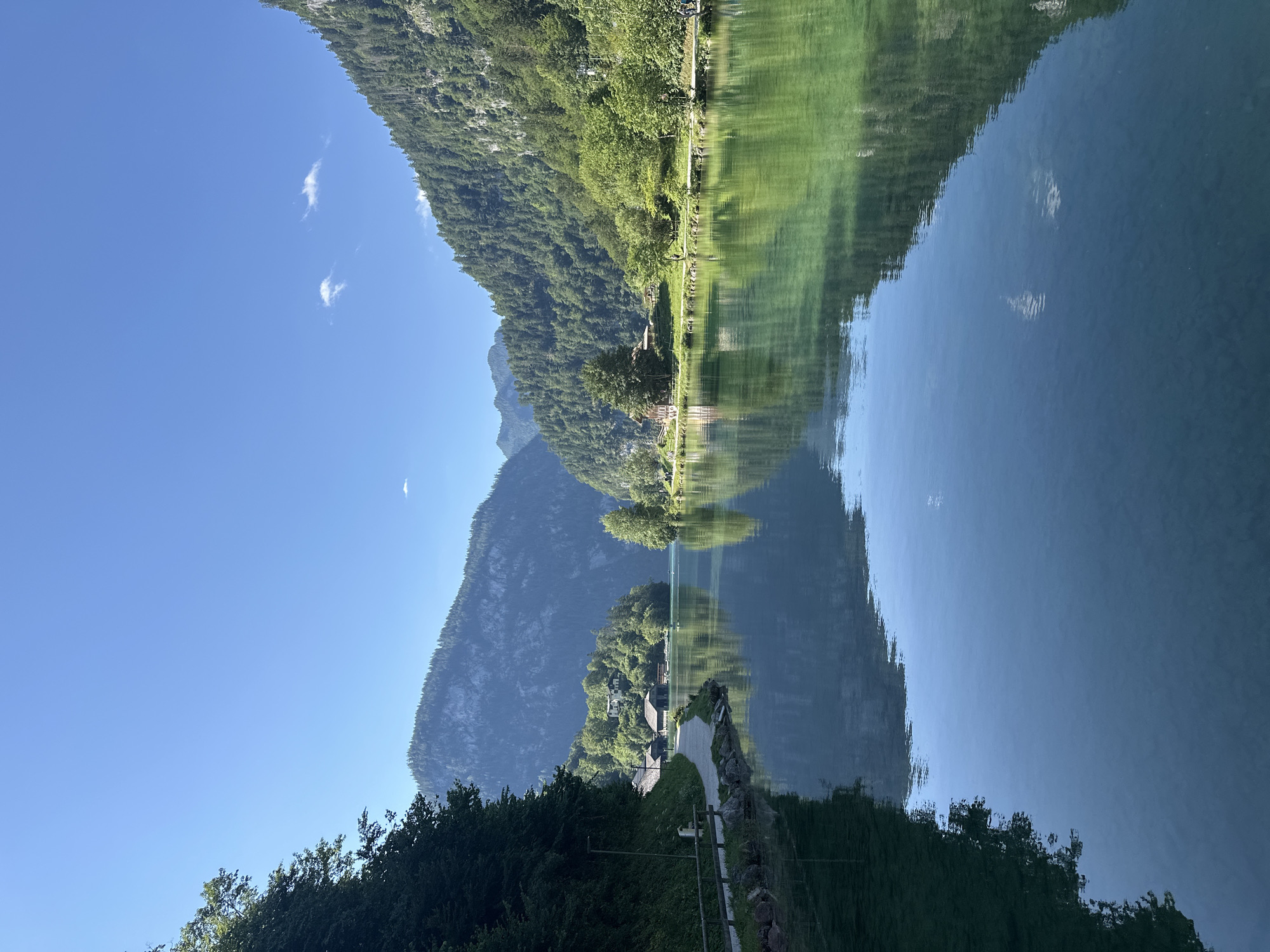 The image depicts a tranquil scene at the Königssee lake in Schönau am Königssee, Germany. A clear blue sky stretches above lush green mountains that flank the serene, reflective body of water. The shoreline features a paved walkway lined with trees and a wooden fence, with a few rustic buildings nestled amongst the greenery. The water's surface acts as a mirror, reflecting the surrounding landscape with striking clarity. The overall impression is one of peaceful natural beauty, with the architecture seamlessly integrated into the environment.