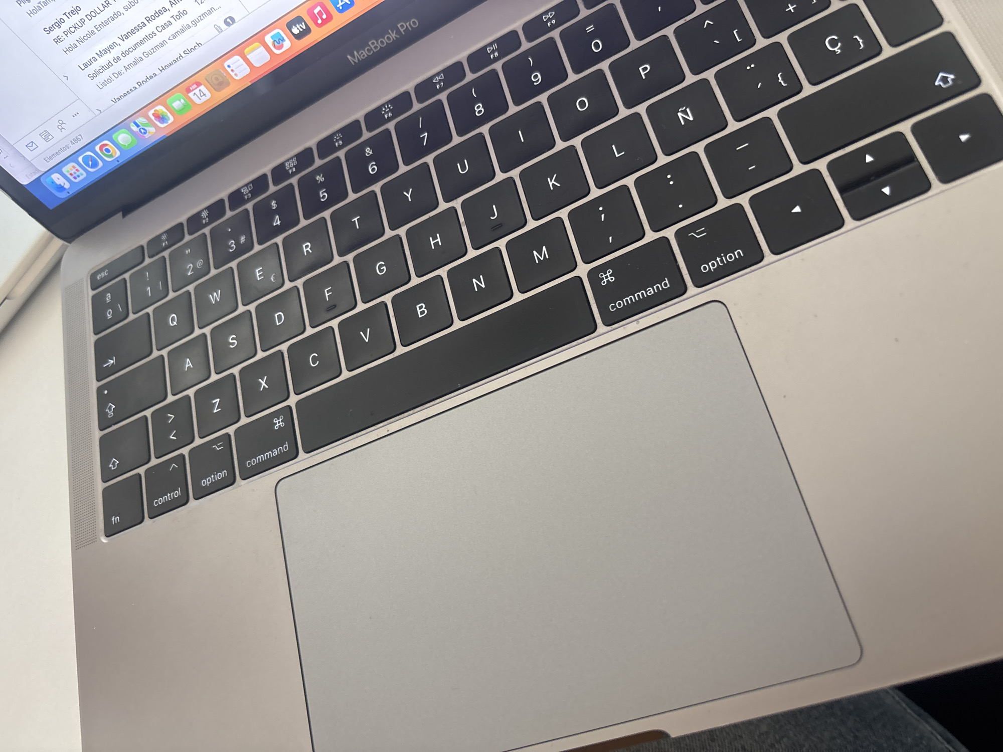 This image is a close-up of a silver MacBook Pro keyboard and part of the screen. The laptop is open, and the screen shows a partially visible email inbox with several email subject lines. The keyboard layout is visible, featuring a Spanish language configuration including the Ñ key. Keys such as "command," "option," "control," "fn," and the arrow keys are clearly shown. The trackpad is also visible. The laptop appears to be resting on someone's lap, and blue jeans are visible along the bottom edge.