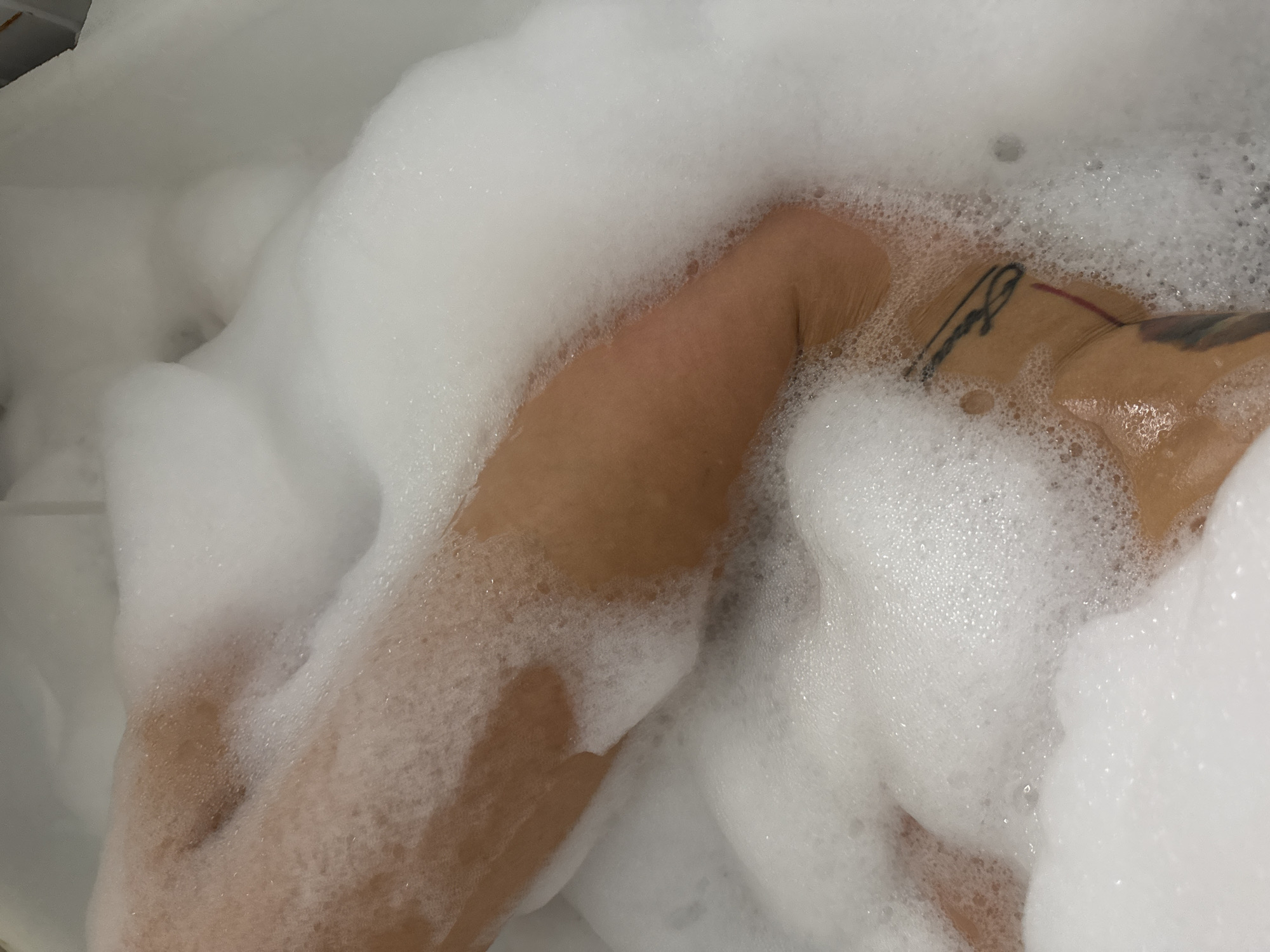 The image depicts a close-up view of a person taking a bubble bath in an indoor setting, likely in Chaikovsky, Russia. The main subjects are the person's legs and lower torso, partially submerged and heavily covered by white, frothy bubbles. The person's skin tone appears to be light to medium brown, with parts of their body visible through the foam.
A notable detail is a tattoo on the upper thigh or hip area, visible through the bubbles. It appears to be a black line-drawing design, possibly script or an abstract pattern, with a faint red line also present near it. The abundance of bubbles suggests a relaxing and comfortable activity. The background consists of white edges, likely the rim or side of a bathtub. The lighting is soft and even, typical of an indoor bathroom environment. No discernible text, other than the ambiguous tattoo, is visible. The overall scene conveys a sense of personal care and tranquility.