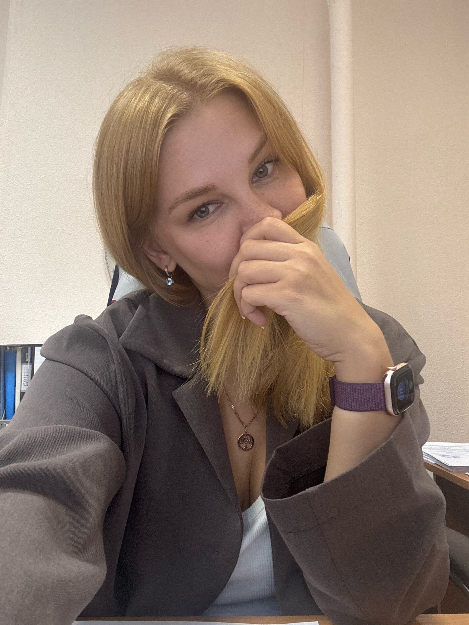 A woman with blonde hair is sitting at a desk, holding a strand of her hair up to her face. She is wearing a grey blazer and a white top, and a smartwatch on her left wrist. The background appears to be an office setting, with shelves and papers visible. The image is taken from a close-up perspective, focusing on the woman's face and upper body. The location of Rusanovo, Russia is not depicted in this image.