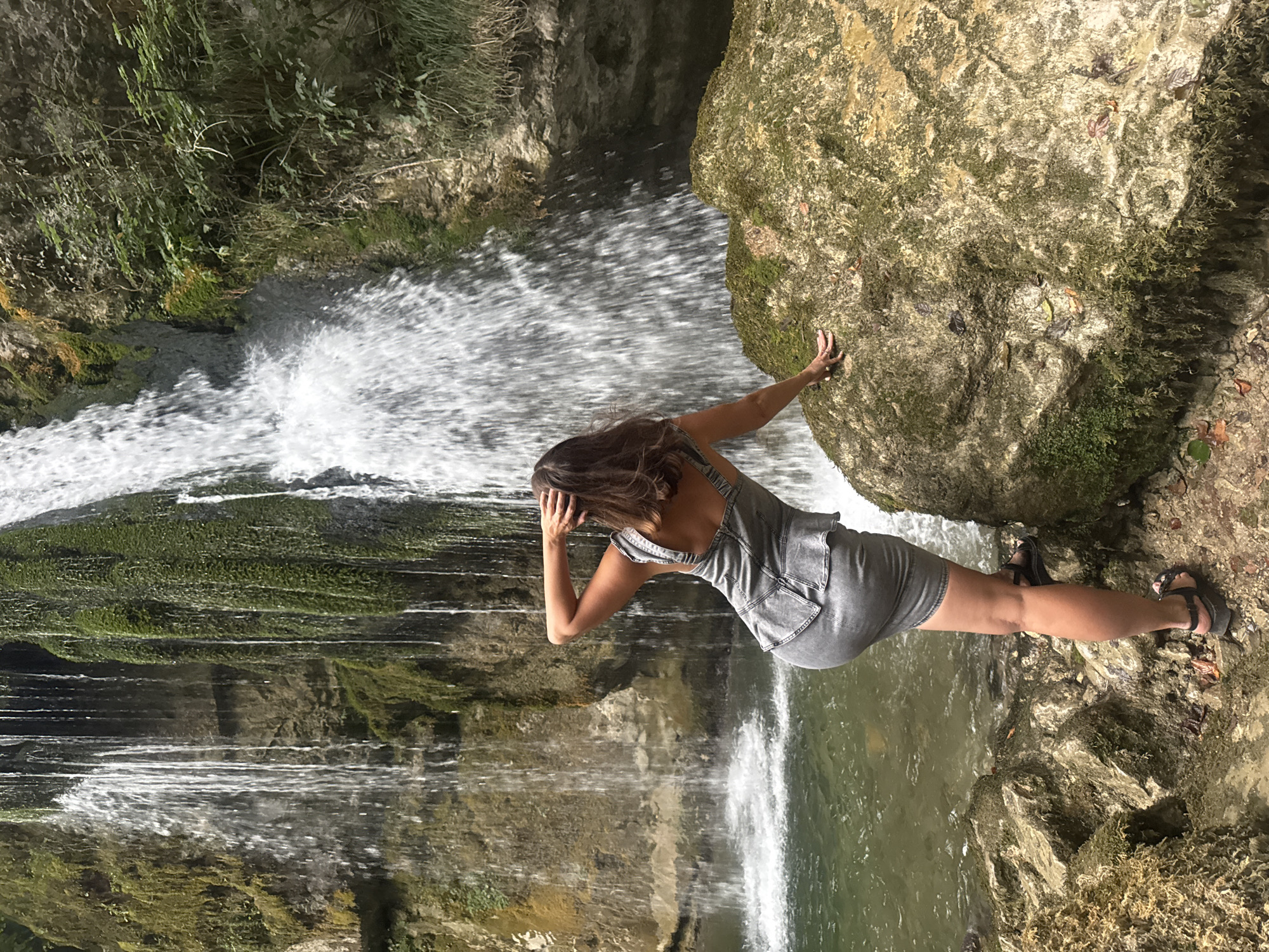 A woman is standing by a waterfall with her back to the camera. She is wearing a denim dress and sandals. Her right arm is raised, and her hand is resting on her head. She is leaning on a large rock covered in moss. The waterfall is cascading down a rocky cliff face. The water is clear, and the surrounding area is lush and green. The location appears to be a natural outdoor setting, possibly a park or a forest area.