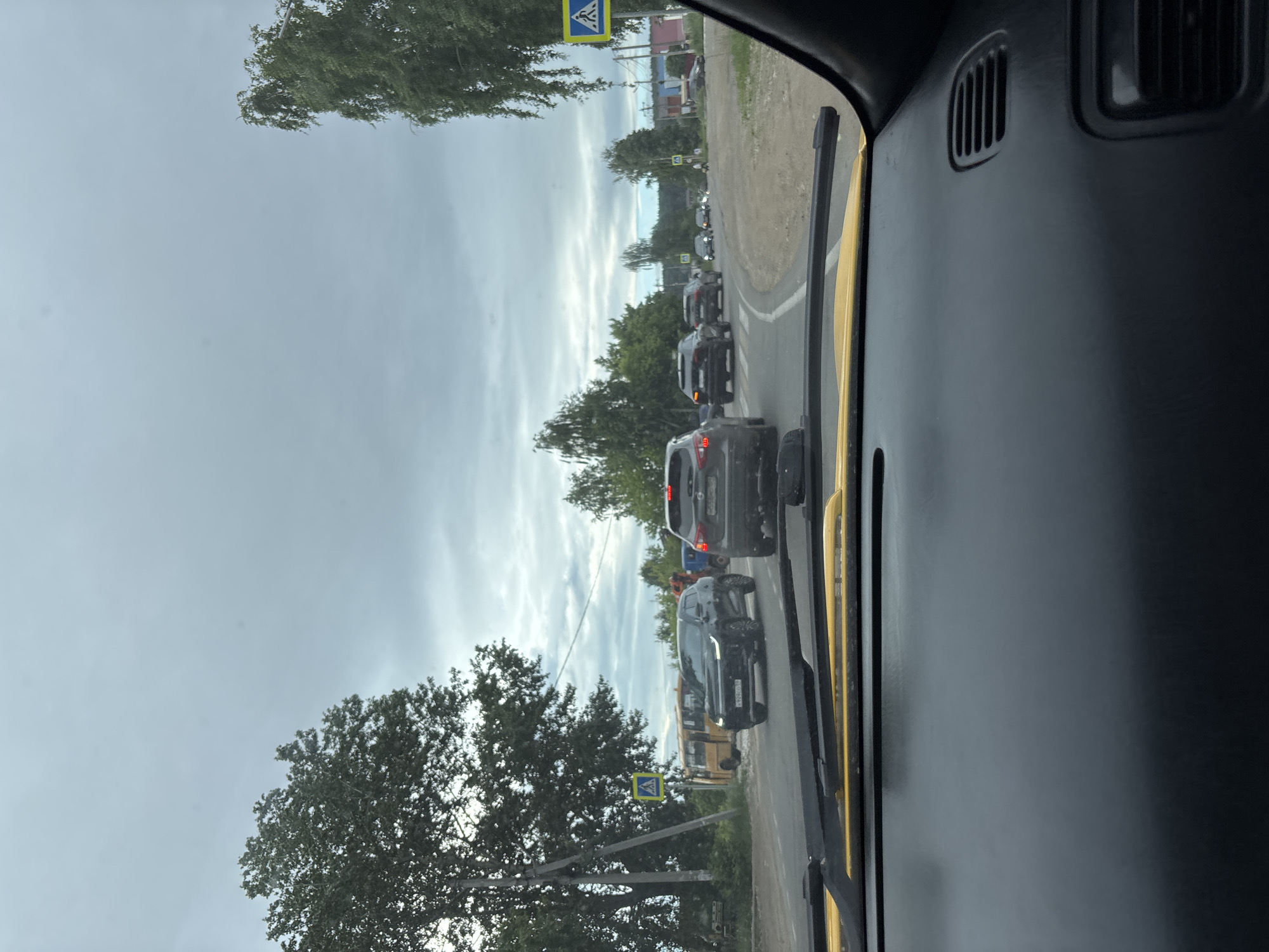 The image shows a view from inside a car, looking out at a traffic jam on a cloudy day. The road is paved and appears to be a rural or suburban area. Several cars are visible, including SUVs and sedans, stopped in traffic. A yellow bus is parked on the side of the road, and a pedestrian crossing sign is visible. Trees line the sides of the road, and the sky is overcast. The dashboard of the car is visible in the foreground, with the windshield wipers resting on the windshield. The location is identified as Chaikovsky, Russia.