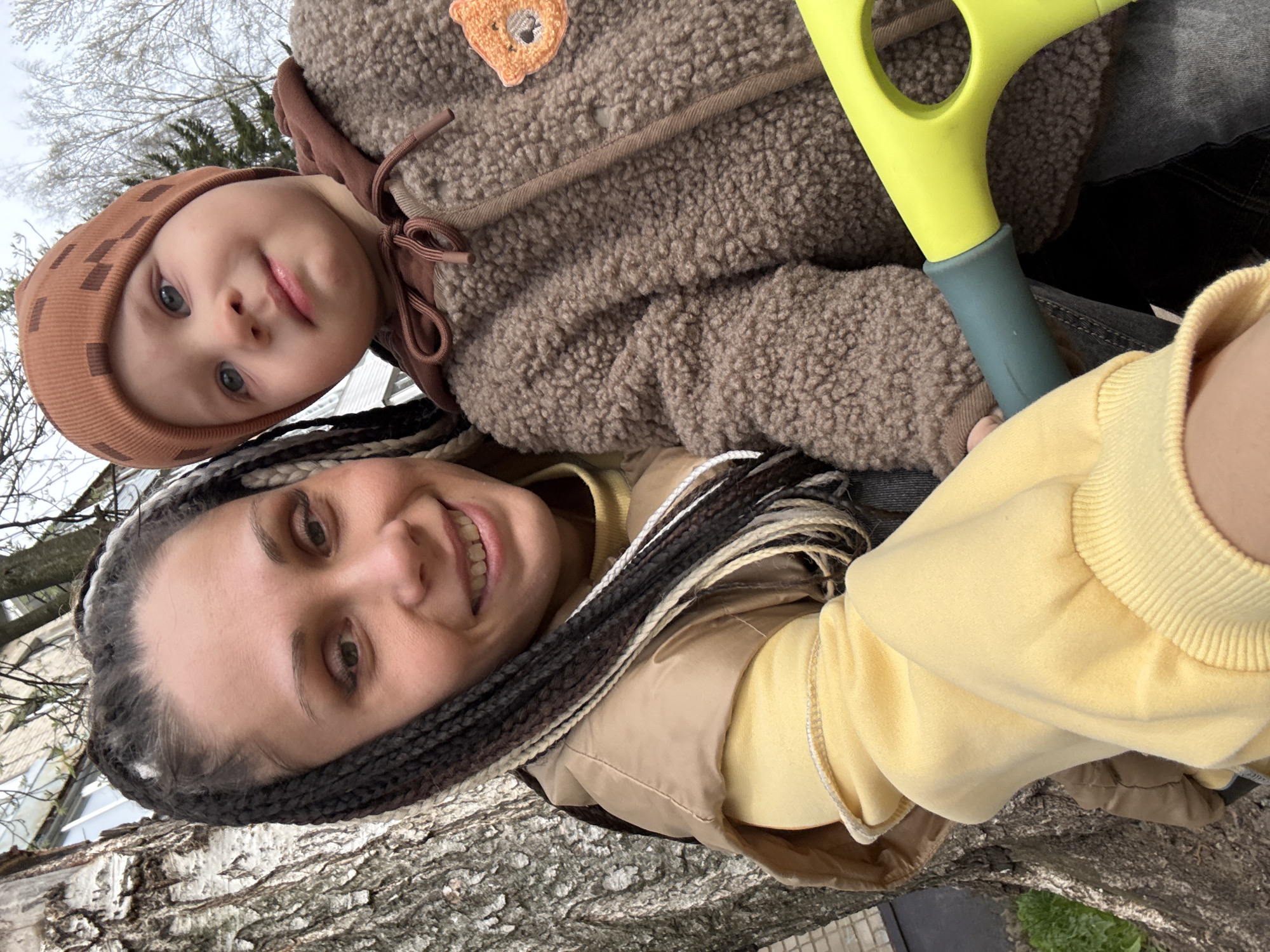 A close-up, low-angle photo captures a woman and a young child outdoors. The image is taken from a perspective looking up, with the background consisting of an overcast sky and bare tree branches. The overall impression is a casual, happy moment between what appears to be a mother and child in Rusanovo, Russia.
In the foreground, a woman is smiling broadly, her face angled slightly upwards. She has dark hair styled in braids or dreadlocks, featuring strands of varying shades including black, white, and brown. She wears a yellow long-sleeved top and a light brown or tan vest or jacket.
Above and slightly behind the woman is a young child, likely a toddler or preschooler, who looks directly at the camera with striking blue eyes and a calm, curious expression. The child is dressed in a warm, patterned brown knitted hat that covers the ears, and a thick, fuzzy brown jacket with a small, light orange teddy bear patch on the upper right side. The jacket has visible drawstrings around the neck.
A bright neon yellow plastic toy, with a blue-grey handle section, is prominent between the child and the woman, possibly being held by the child. The toy appears to be part of a shovel or similar play equipment.
The immediate foreground features the textured, rough bark of a tree trunk, possibly a birch, indicating their proximity to trees. In the background, beyond the bare branches, parts of a light-colored building are visible, suggesting an urban or suburban setting. The overcast sky indicates daytime, and the warm clothing suggests cool weather, possibly early spring or late autumn. There is no visible text in the image.