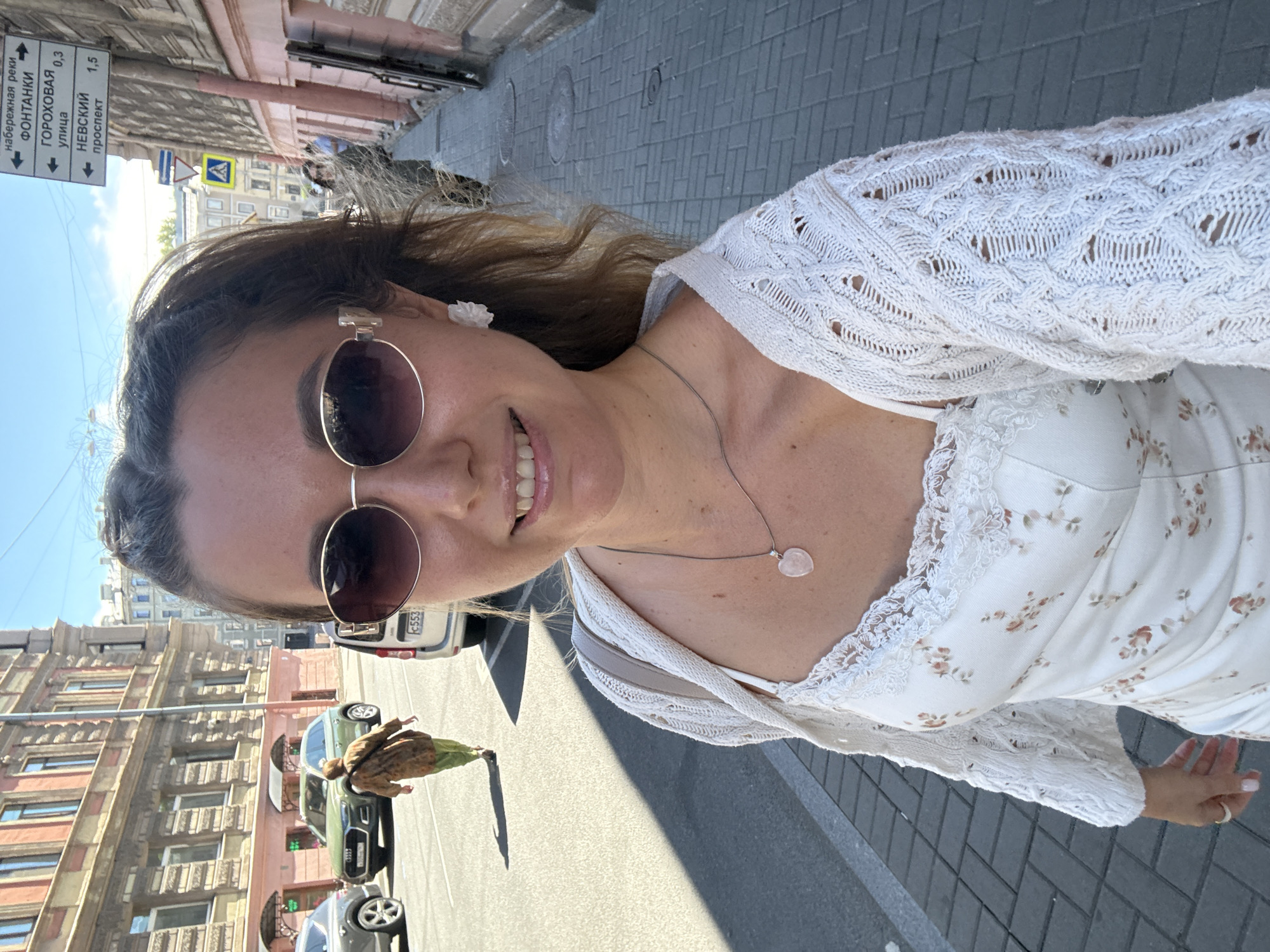 The image is a well-lit, eye-level outdoor selfie featuring a woman on a paved city sidewalk. The location is in the city of Unknown, Unknown. The woman, positioned centrally, is smiling directly at the camera. She has medium-long, wavy brown hair, partially pulled back, and wears large, oval-shaped sunglasses with dark lenses and light-colored frames. Her lips are slightly parted, revealing upper teeth, and appear glossy. A white, flower-shaped stud earring is visible on her right ear (viewer's left). She wears a delicate silver-colored chain necklace with a light pink, heart-shaped pendant. Her attire consists of a white dress with a subtle floral pattern in reddish-brown and green hues, featuring thin straps and a lace trim along the neckline. Over this, she wears an open-front, long-sleeved white cardigan with intricate cable and eyelet knit patterns. Her right hand (viewer's left) is visible, showing light-colored nail polish and a simple ring on the ring finger. A light beige or tan shoulder bag strap is visible over her left shoulder (viewer's right). To her right, the sidewalk is paved with grey rectangular pavers. Several round metal utility covers are embedded in the sidewalk. A tall, dark metal pole stands on the edge of the sidewalk, displaying a white rectangular street sign with black text and arrows in Cyrillic. The sign indicates directions to "набережная реки ФОНТАНКИ" (Fontanka River Embankment) at 0.3 km, "ГОРОХОВАЯ улица" (Gorokhovaya Street) at 0.3 km, and "НЕВСКИЙ проспект" (Nevsky Prospekt) at 1.5 km. Below this, a blue rectangular sign with a red diagonal line (indicating no stopping/parking) and a yellow diamond-shaped sign with a black pedestrian symbol (indicating a pedestrian crossing ahead) are affixed to the pole. To the woman's left is an asphalt road with visible white lane markings. Direct sunlight casts prominent shadows across the road surface. A grey SUV is partially visible on the far left. Further down the street, a dark green Audi SUV (possibly Q5 model) is seen, followed by an older woman walking away from the camera, dressed in a brown patterned top and green pants, carrying a dark shoulder bag. A silver van with a visible license plate "C553" is partially obscured by the woman on the right side of the road. The background features multi-story city buildings under a clear blue sky with a few wispy white clouds. The building on the left side of the street has reddish-pink lower