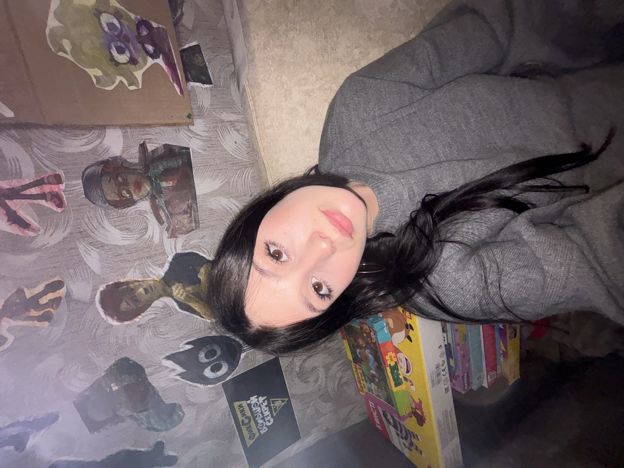 An indoor close-up photograph depicts a young female subject in a domestic setting, located in the city of Фоки, Russia. The subject has long dark hair, dark eyes with visible eyelashes, and light pink lips. She is wearing a grey knitted crew-neck sweater and a small hoop earring is visible in her left ear. Her head and upper torso are visible, and she looks directly at the camera with a neutral expression.
Behind the subject, a wall is covered in light-colored wallpaper featuring a subtle swirling or patterned texture. Multiple paper cut-out figures are affixed to the wall. These include a black character with large white eyes and a flame-like top, positioned above a black rectangular sign with white Cyrillic text reading "ФИКСИКИ БОЛЬШОЙ СЕКРЕТ" (Fixiki The Big Secret). Other cut-outs depict a figure in a helmet with red lips, an abstract cartoon-like face on cardboard, and various partial or indistinct forms.
To the subject's right and behind her, a light beige upholstered surface, consistent with a couch or bed headboard, is visible. To her left, in the lower foreground, a stack of colorful cardboard boxes is present. The top box displays illustrations of cartoon animals and Cyrillic text, including "СУДОКУ 160 ЗАДАНИЙ" (Sudoku 160 Tasks). The scene is illuminated by artificial light from the front, casting soft shadows.
