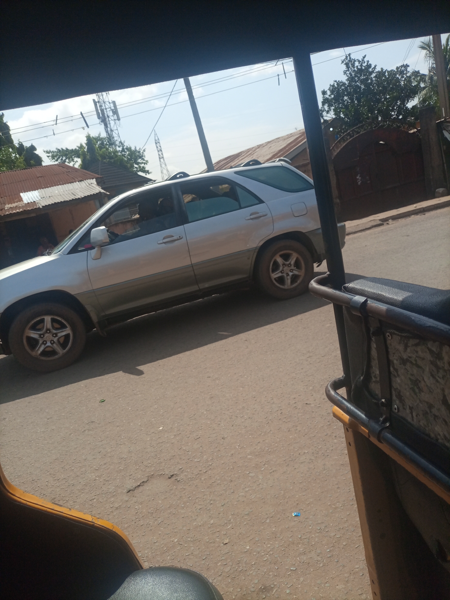 This media file shows a street scene in Nkpor, Nigeria, on a bright, sunny day. The perspective is from inside a three-wheeled vehicle, likely a commercial motorcycle or "okada" commonly used for transportation in Nigeria. In the foreground, the yellow and black seating and handlebars of the vehicle are visible.
The main subject of the image is a silver SUV, possibly a Lexus RX, driving on a paved road. The SUV is slightly dusty, indicating it has been driven on unpaved surfaces or in a dusty environment. The vehicle's tires are also dusty. The SUV has tinted windows, and a person can be seen driving it.
In the background, there are several buildings with corrugated metal roofs, typical of residential or commercial structures in the area. A large metal gate, possibly an entrance to a property, is visible on the right. A tall cell tower stands prominently in the distance, suggesting the presence of modern communication infrastructure within the otherwise seemingly traditional setting. Green trees and foliage are scattered throughout the scene, adding to the natural environment. The sky is clear and blue, confirming the sunny weather. The overall impression is a snapshot of daily life and transportation in a Nigerian town. No visible text is present in the image.