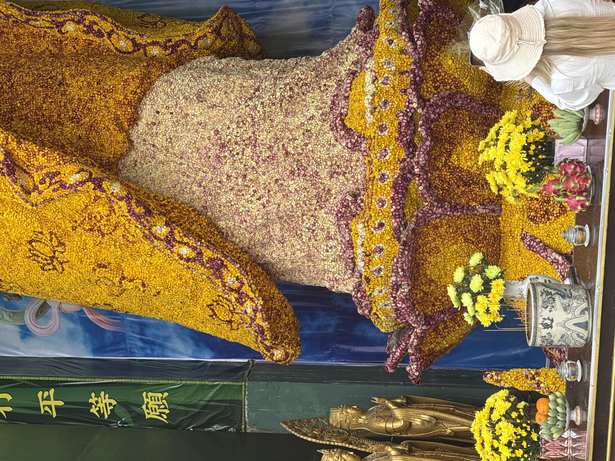 A high-angle, indoor photograph captures an elaborate floral display, likely for religious purposes, in what appears to be Xuân Thành, Vietnam. The floral arrangement, predominantly yellow and beige with purple accents, is likely a religious offering. A fair-skinned woman wearing a cream-colored hat and white shirt leans over the display, holding a mobile phone, perhaps taking a picture. Alongside the floral structure are arrangements of yellow flowers, dragon fruit, bananas, and other offerings placed on a shelf. Several small, decorative containers are also present. Two golden statues of Buddha are seen below the shelf. A banner with green background and yellow Chinese characters adorns the space beneath the shelf. A backdrop behind the flower display, blue in color, adds depth to the scene.