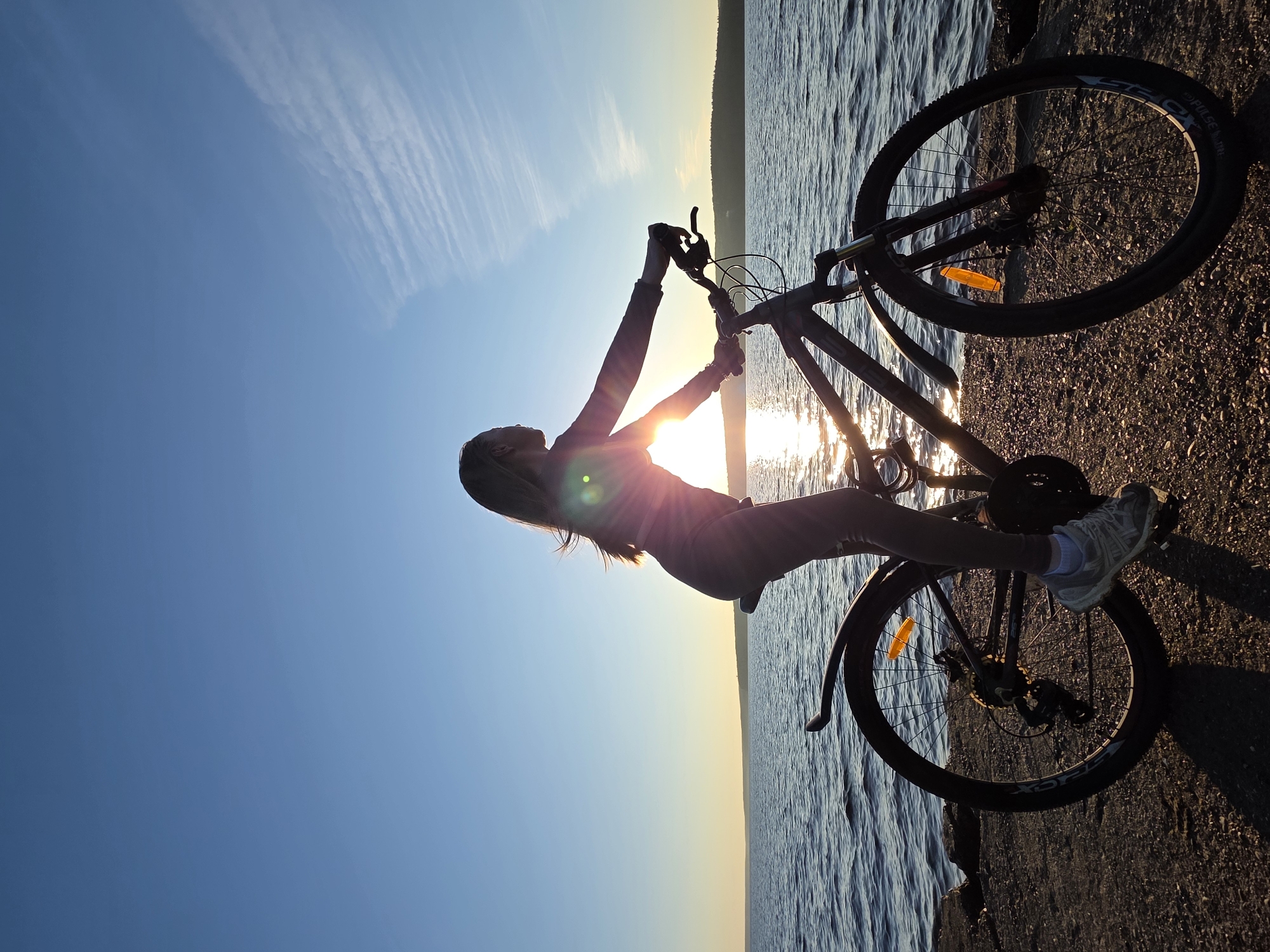 The image shows a silhouette of a woman on a bicycle next to a body of water, possibly the Kama River, in Chaikovsky, Russia. The sun is setting, casting a warm glow on the scene. The woman is wearing athletic clothing and appears to be enjoying the view. The bicycle is a mountain bike with disk brakes. The ground is covered in gravel and rocks. The water is calm with gentle ripples. The sky is a clear blue with a few wispy clouds. The overall atmosphere is peaceful and serene.