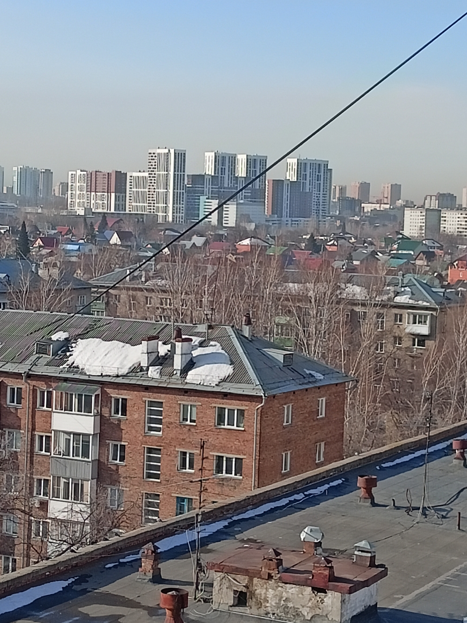 A high-angle view captures a cityscape, with a foreground occupied by the flat rooftop of a building constructed from concrete, showing signs of decay and age. On the roof is an array of infrastructure, including ventilation systems, chimneys, and rusted metal components.
Beyond the immediate rooftop lies a four-story brick building with a grey, gabled roof partially covered in melting snow. The brick building is built in the style of the early Soviet Union period, with straight lines, and minimalist design.
Further into the midground is a mix of single-family houses with brightly-colored roofs. Bare trees populate the spaces between the buildings, indicating the winter season.
In the background, the skyline of Novosibirsk is visible, dominated by tall, modern apartment buildings. The sky overhead is pale blue, hazy with smog. A black electrical cable stretches diagonally across the frame.