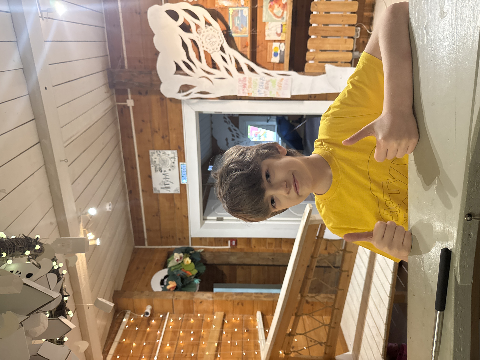 A photo shows a young person with brown hair wearing a yellow shirt, positioned at a table with a grey surface. They are giving a thumbs-up sign with both hands. The room is wood-paneled with decorative elements including a large white snowflake cut-out above a doorway, string lights, and floral arrangements. A sign with the word "Moscow" is visible in Russian near the doorway. Other artwork and decorations adorn the walls. A ladder leans against the wall behind the person.