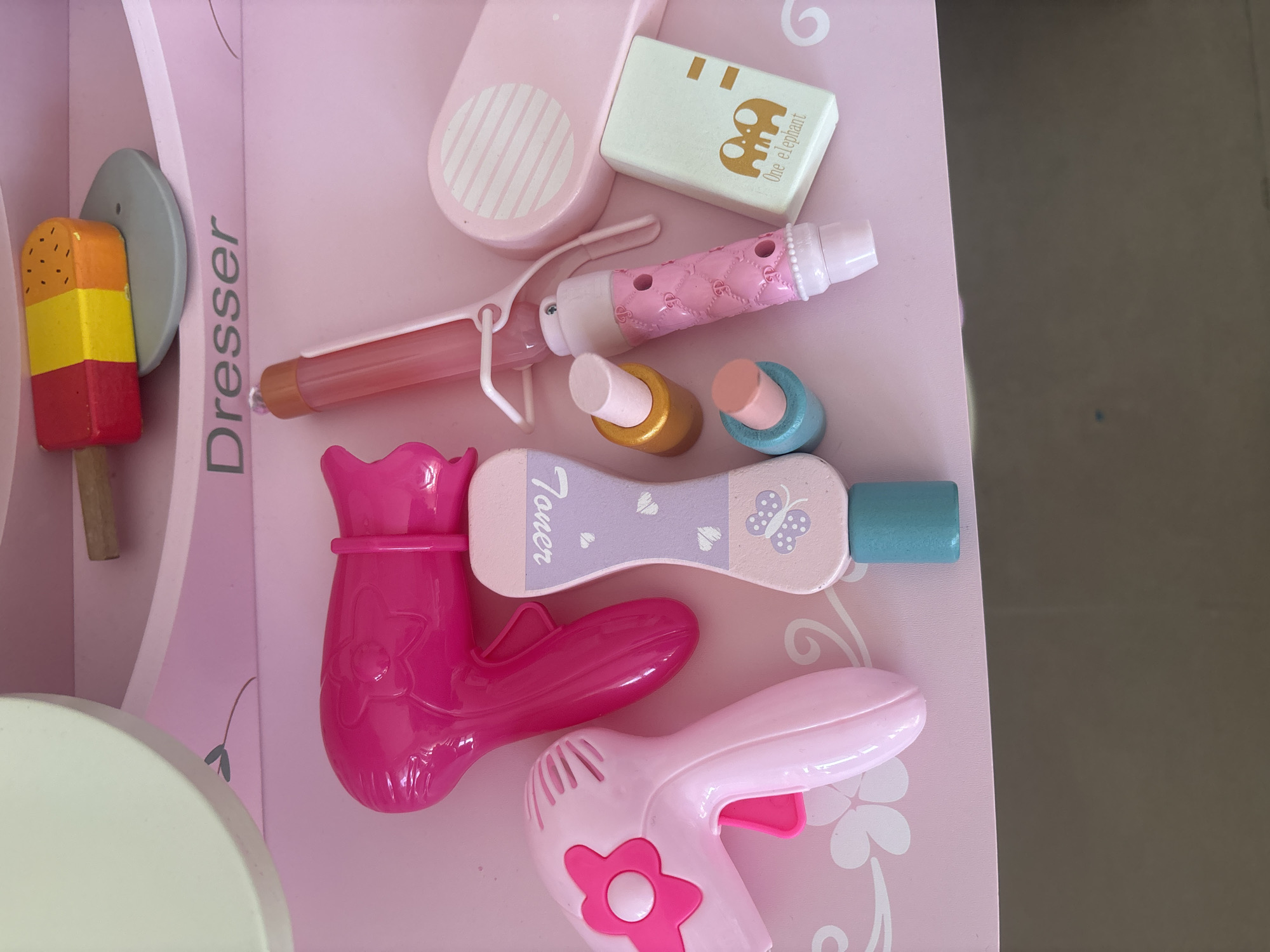 The image shows a collection of toy beauty products, including hairdryers, a curling iron, nail polish bottles, a makeup compact, and a wooden popsicle. The items are arranged on a pink dresser, with the word "Dresser" printed in gray lettering above them. The dresser is part of a larger toy set, with a round white element visible in the top left corner, likely representing a mirror or vanity top. The overall aesthetic is playful and geared towards children. The scene is set in a domestic indoor environment, with a dark, out-of-focus surface visible at the bottom of the frame, suggesting the toys are on a table or floor. There are no people present in the image. The lighting is bright and even, typical of an indoor setting. There is no information in the image to suggest a specific location in Dubai, United Arab Emirates; the image depicts a generic toy set.